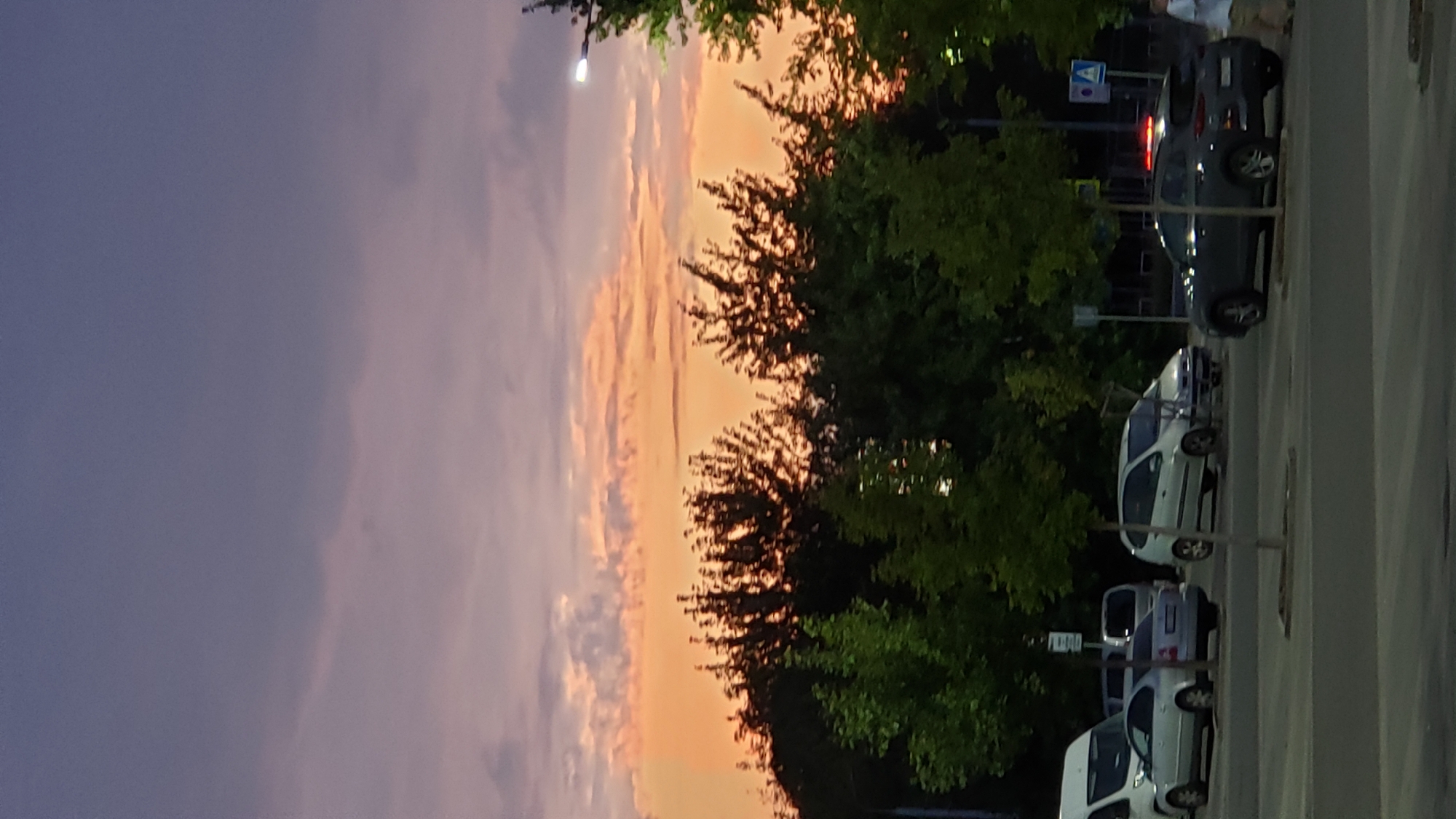 The image captures a dusk scene in Prešov, Slovakia, featuring a parking lot with several cars and trees silhouetted against a vibrant sky. The sky displays a gradient of colors, from a deep purple at the top to shades of orange and pink near the horizon, indicating the setting sun. Clouds are visible, tinged with the warm colors of twilight.
In the foreground, a paved parking area is visible with several vehicles parked. These include a white van on the far left, a silver sedan next to it, and a light-colored hatchback further to the right. A darker-colored SUV with red brake lights is positioned in the middle right of the frame. Two pedestrians are partially visible on the far right, one walking away from the camera.
Behind the parking lot, a line of trees forms a dense backdrop. The trees vary in height and foliage, with some appearing greener and closer, while others are darker and more distant, their branches starkly outlined against the colorful sky. Streetlights and traffic signs are sparsely present within the scene. The overall atmosphere is calm, with the natural beauty of the sunset contrasting with the urban setting of the parking lot.