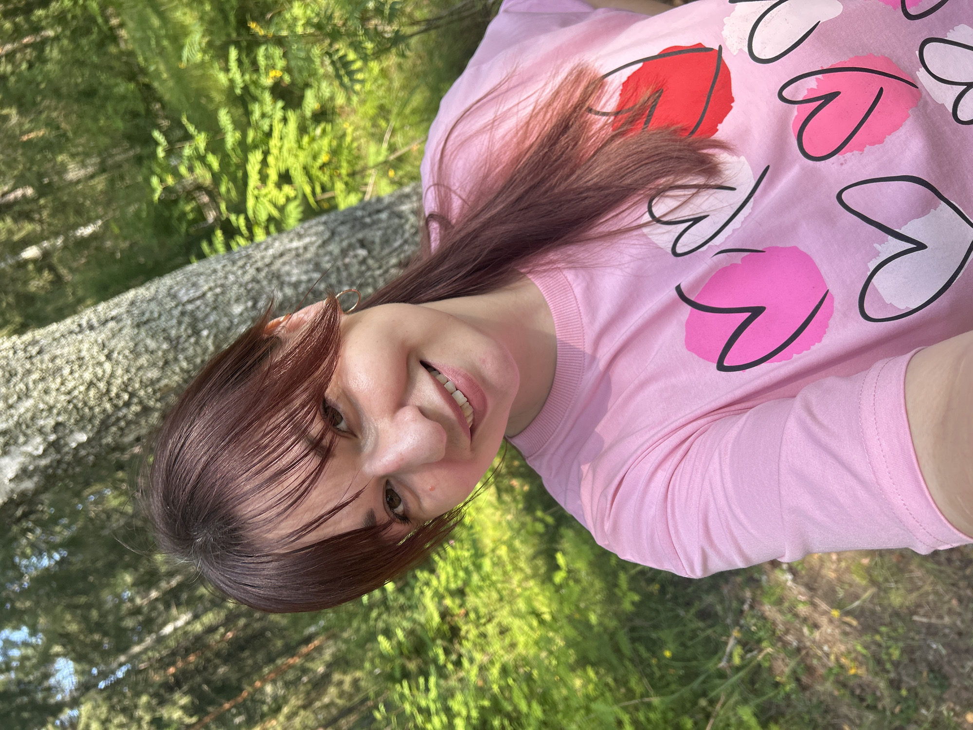 A young woman with brown hair and bangs is taking a selfie in a forest. She is smiling and wearing a pink t-shirt with various hearts printed on it. The background shows a birch tree and lush green foliage, indicating an outdoor, natural setting. The lighting suggests it is daytime, likely sunny, given the bright illumination of the scene. The overall impression is casual and relaxed. The location is the city of Krasnovishersk, Russia.