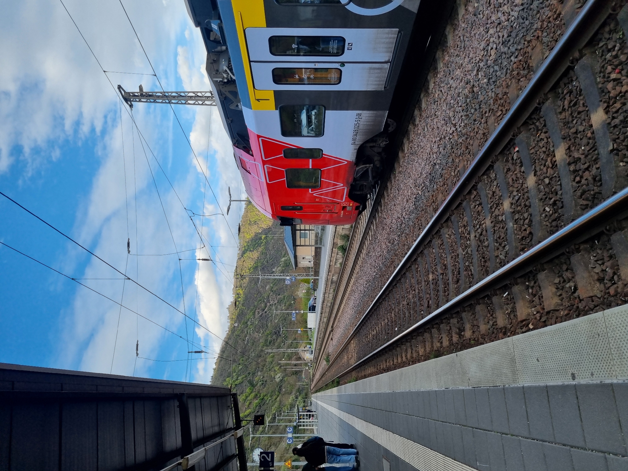 The image shows a train stopped at a platform in Cochem, Germany. The train is red, yellow, and gray, with the number 94 80 34624 025-5 D-DB visible on the side. The platform is gray and tiled, with a black metal structure overhead. A person is seated on the platform near a suitcase. In the background is a hillside covered with trees, with buildings visible further back. The sky is blue with white clouds. Power lines and overhead supports are also visible above the train. The railway tracks run parallel to the platform.