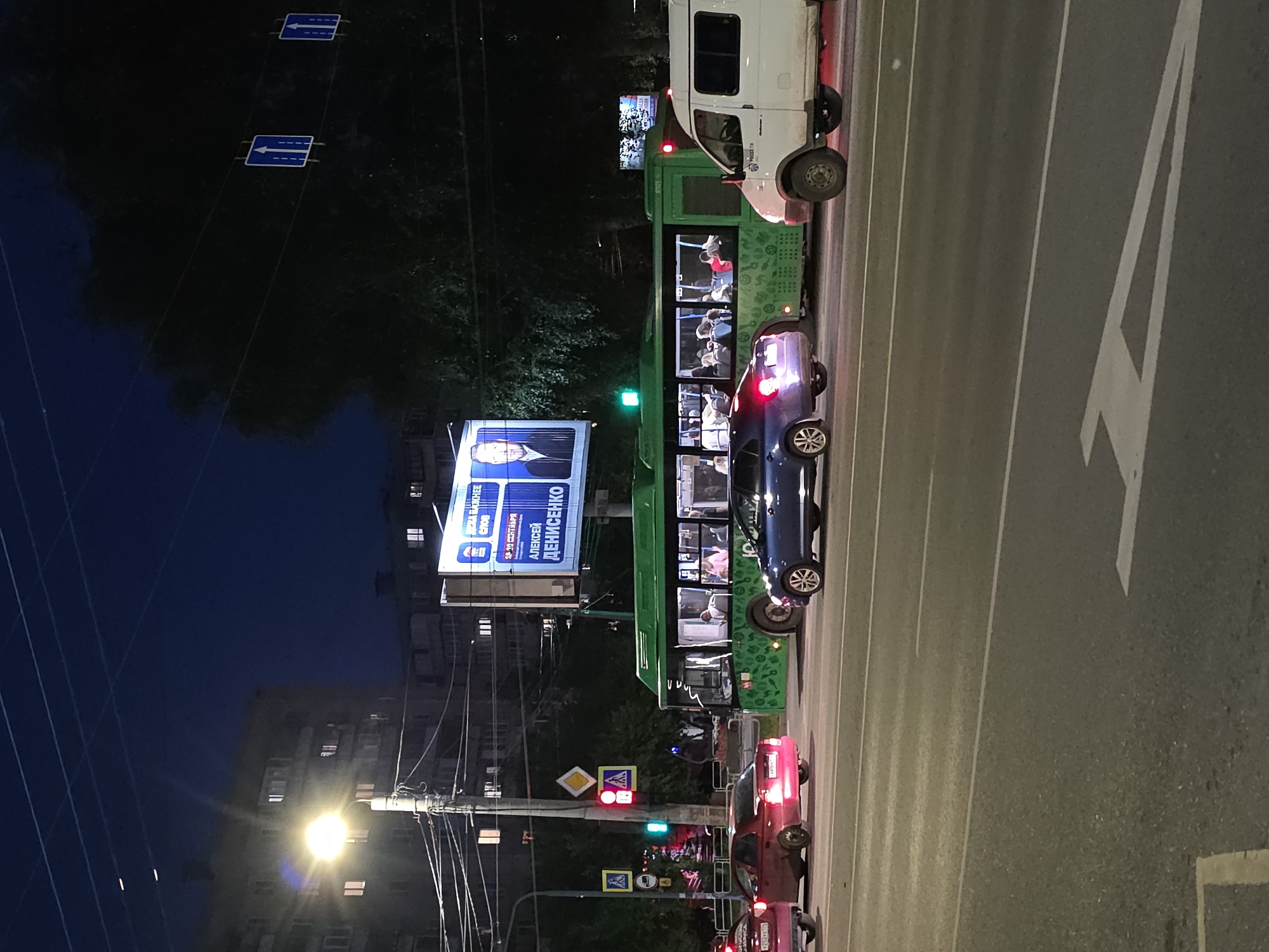 This is a nighttime, eye-level, medium shot of a street scene in Chelyabinsk, Russia. The image captures a busy intersection with traffic, a large green public bus, a white van, and several cars. A billboard featuring a man's face and Russian text is prominently displayed in the background. Traffic lights, streetlights, and the lights from buildings and vehicles illuminate the scene. The sky is dark blue, indicative of late evening or early morning. The road is asphalt with visible lane markings. The overall impression is of urban nighttime activity.