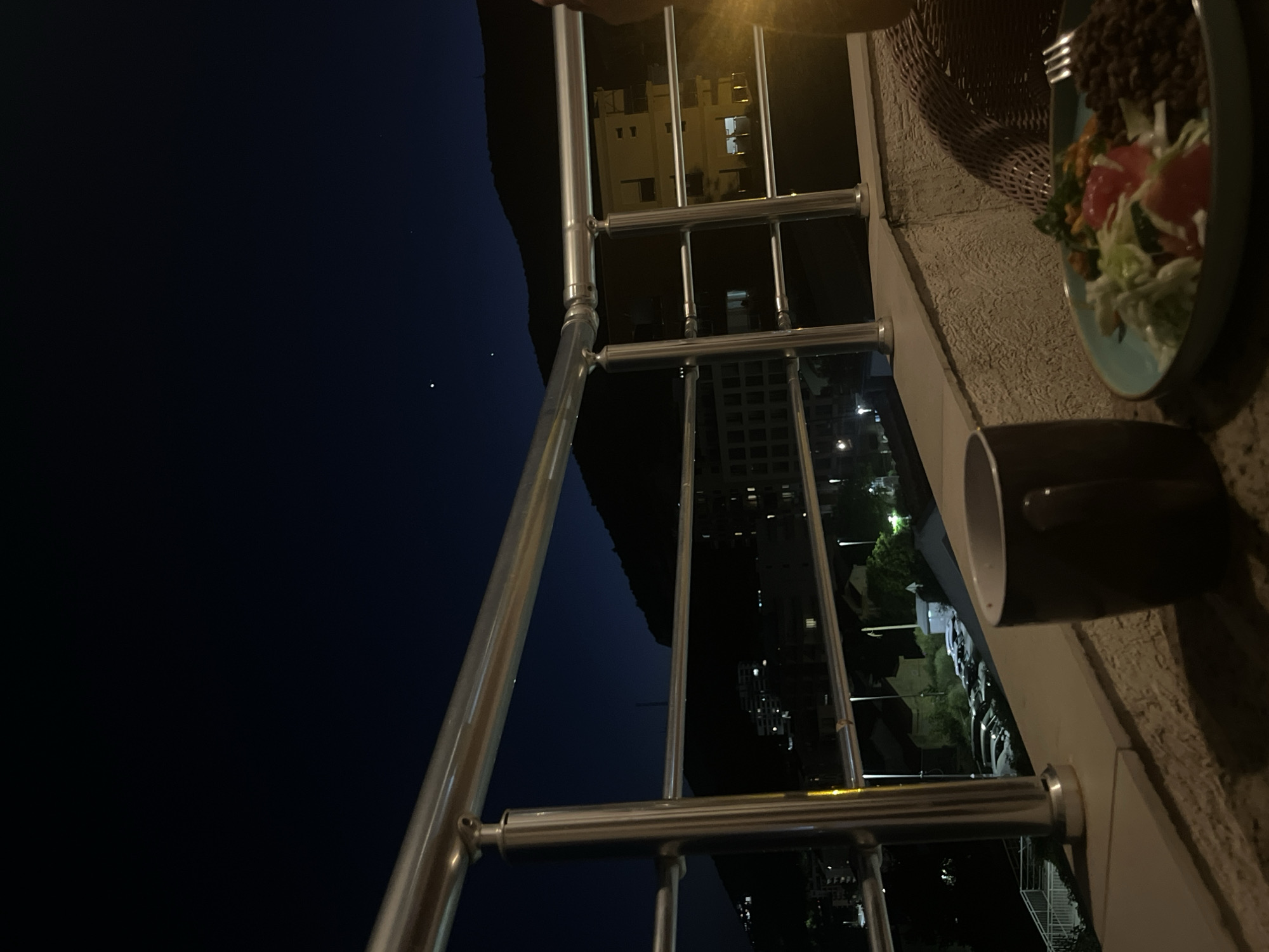 The image depicts a balcony at night, overlooking a city and a hillside. A metal railing runs across the foreground, with a dark mug and a plate of food on the balcony's edge. The plate contains a salad with lettuce and tomatoes, and a side of dark, granular food, possibly lentils or beans. A fork rests on the right side of the plate. In the background, city lights are visible, along with buildings, parked cars, and streetlights. A large hill or mountain forms the backdrop, with a few distant lights suggesting structures on its slopes. The sky above is dark blue, with a few faint stars or celestial bodies visible. The scene is set in the city of Bečići, Montenegro.