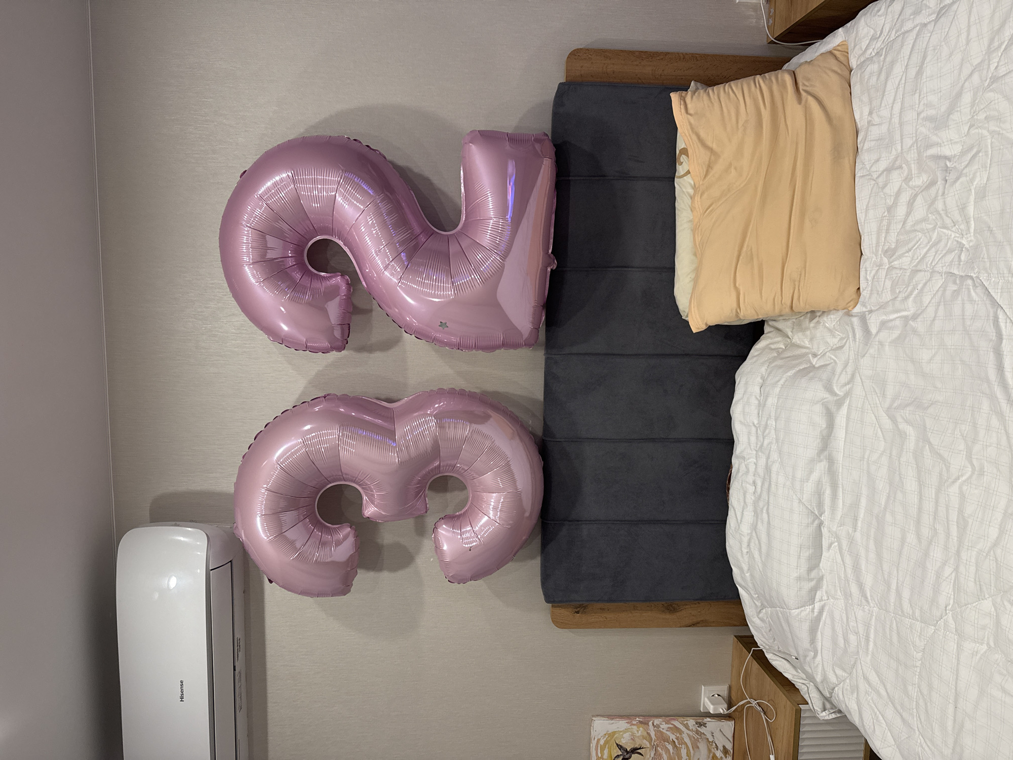 The image shows a bedroom. Pink foil balloons in the shape of the number 23 are affixed to the wall. Next to the balloons is a bed with a grey headboard and a white comforter. A peach-colored pillow is on the bed. An air conditioning unit is mounted on the wall below the balloons. The location is Unknown, Unknown.