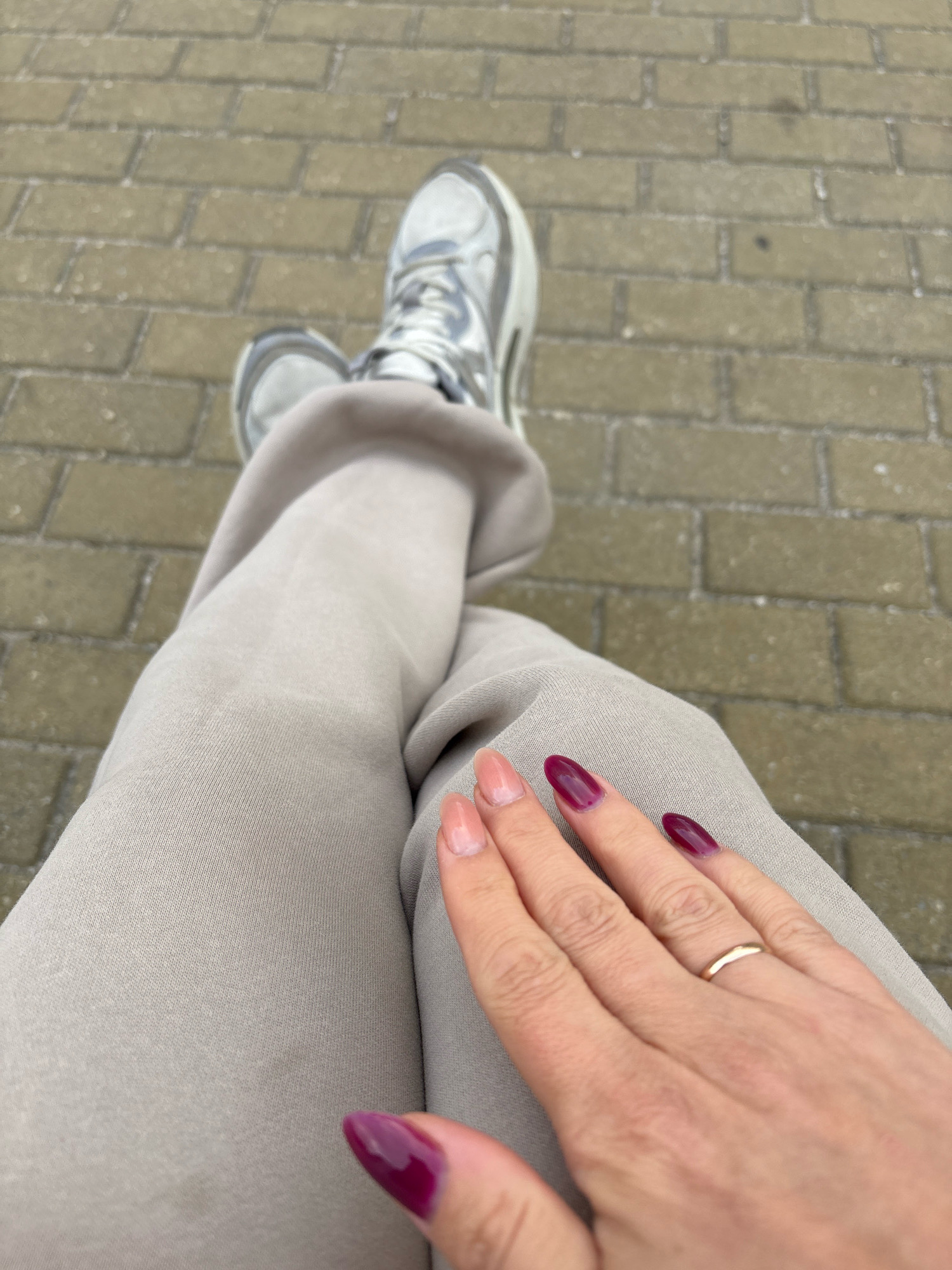 The image shows a person's legs and hands from a high-angle perspective. The person is wearing light-colored pants and silver sneakers, with their legs crossed. The background consists of a paved walkway made of rectangular bricks. The person's hand is visible in the foreground, with fingernails painted in shades of light pink and deep maroon. A thin gold ring is worn on the ring finger. The lighting suggests it is daytime. The location is not identifiable as Zapadny, Russia, based solely on the visual information provided.