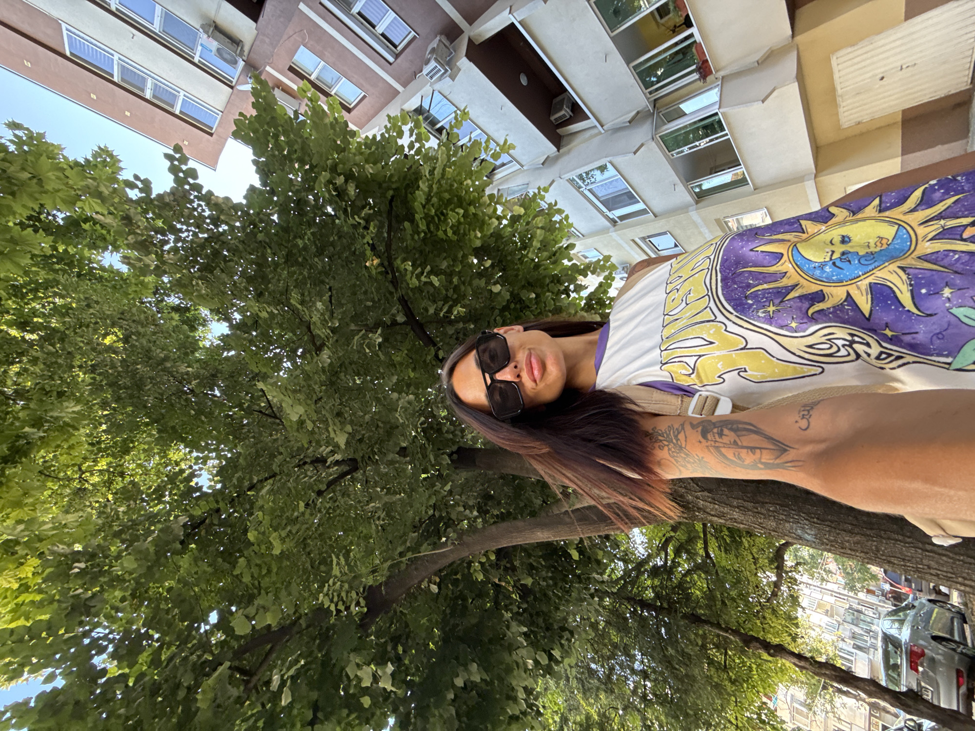 A low-angle, wide-shot, full-body selfie is taken outdoors, capturing a woman standing near a tree and a building in Pleven, Bulgaria. The woman, with dark hair and wearing large sunglasses and a white tank top with a sun and moon graphic, looks towards the camera. Her left arm, adorned with a tattoo of faces and script, is extended, holding the camera. Behind her, a large tree with lush green leaves occupies the left side of the frame, with a multi-story building featuring brown and beige facades and numerous windows on the right. The sky is visible through the tree canopy, and a silver car is parked on the street in the background. The overall scene suggests a sunny day in a city environment.