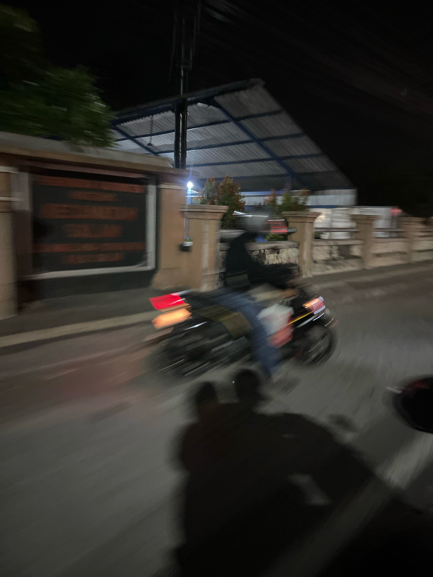 The image captures a nighttime scene on a street in Ngluwar, Indonesia, characterized by motion blur. A person is riding a dark-colored motorcycle, moving from left to right. The motorcycle's taillights are illuminated, appearing red and yellow, and its headlights are also visible. The rider is wearing what appears to be a dark jacket and blue jeans. The environment includes a building with a dark facade and lighter-colored signage on the left, partially obscured by motion blur. In the background, there is a roof structure with a corrugated metal surface and supporting pillars, typical of some Indonesian architecture. Further in the background, there are other buildings and streetlights. The road surface is wet, reflecting the lights. Shadows of the motorcycle and its rider are cast on the road. The overall impression is of a transient moment captured during nighttime movement.