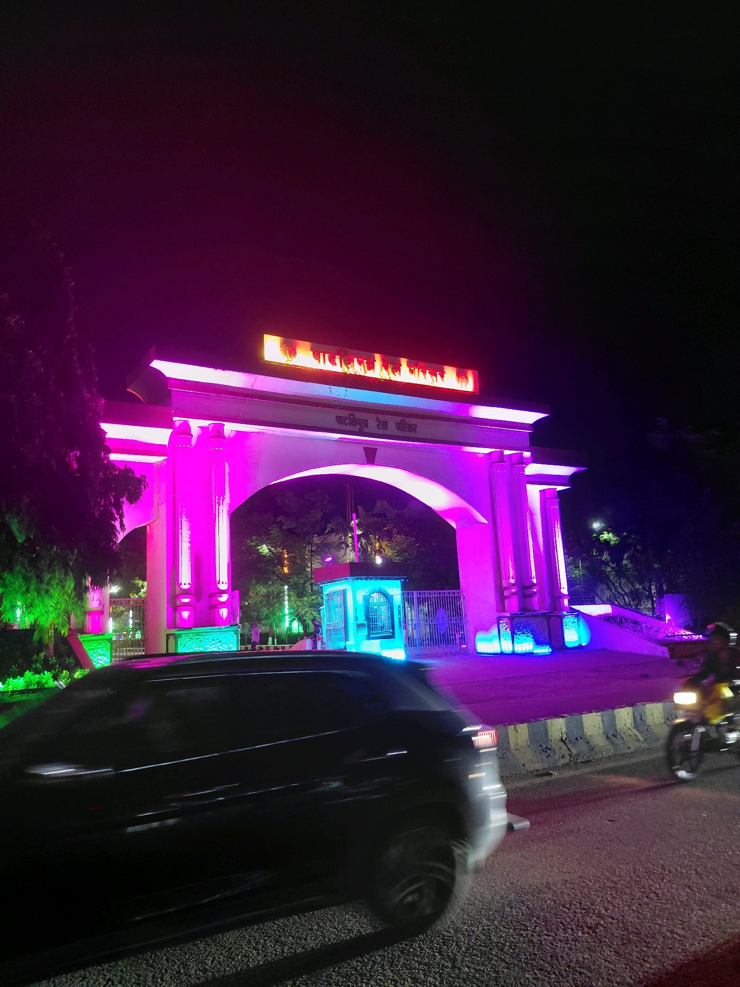 This is a nighttime, eye-level, medium shot of the main entrance archway of the Patna Junction Railway Station in Patna, India, illuminated by vibrant pink and purple lights. A black car is in motion in the foreground, its form blurred due to movement. To the right, a yellow motorcycle with a rider is also in motion, its headlight visible. The archway itself is a substantial structure with pillars and an overhead beam, all bathed in bright pink and purple lighting, with some green lights visible on the left side. Above the arch, there is a lit sign with text in Hindi, and below it, smaller text in Hindi is also visible. Behind the arch, trees and a small, blue-lit structure with a metal gate are discernible. The ground in front of the archway is paved, and a road with a striped curb is in the foreground. The overall atmosphere is festive due to the colorful lighting.