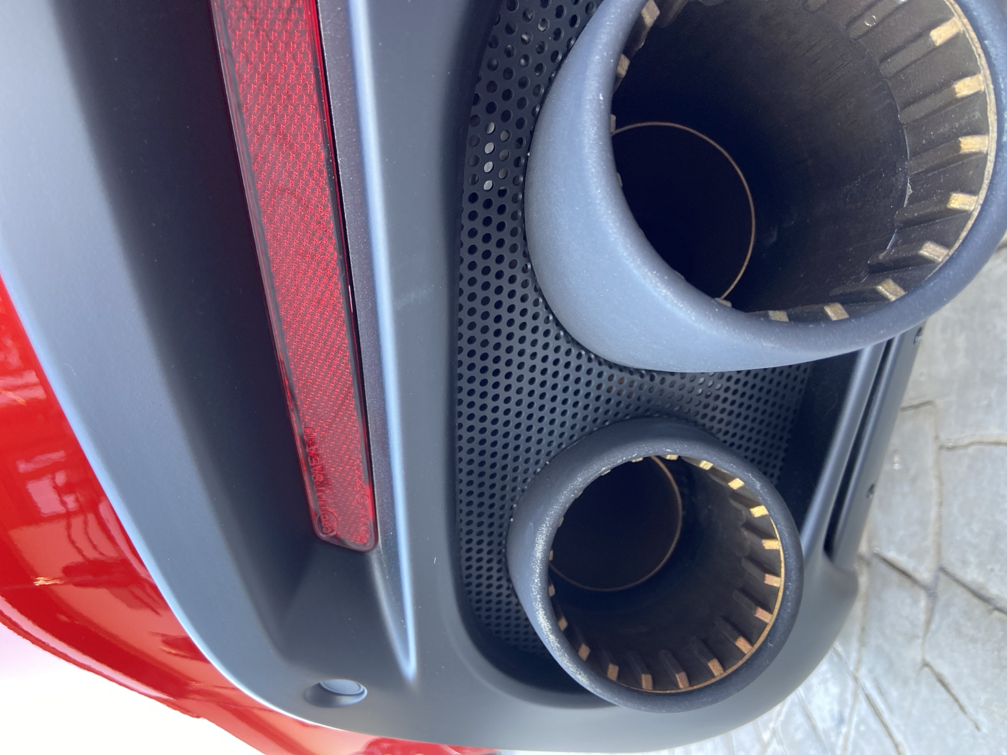 The image shows the rear of a red vehicle, specifically the exhaust pipes and taillight area. The exhaust consists of two circular pipes with grey surrounds, set into a black, perforated panel. The taillight is a vertical red strip. The surrounding bodywork is grey. The vehicle is parked on a paved surface, possibly in a garage or parking area. There is no indication of location or people.