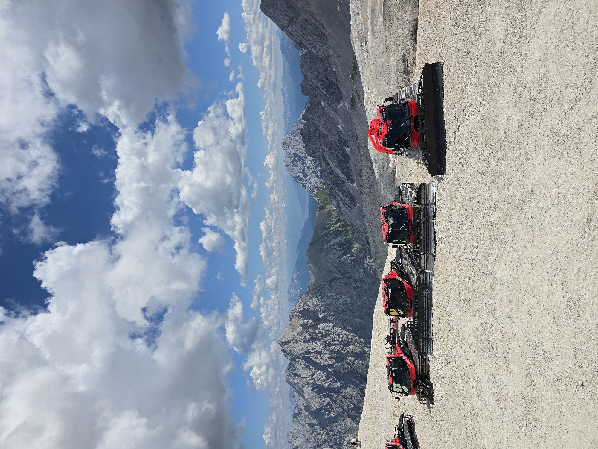 The image depicts a mountainous landscape with several red snow groomers parked on a gravelly surface. In the background, majestic, rugged mountains stretch across the horizon under a bright blue sky filled with scattered white clouds. The scene appears to be a high-altitude location, likely part of a ski resort or mountainous terrain accessible by vehicle. One person is visible in the far left of the image, standing on the edge of a slope. The snow groomers are the primary focus in the foreground, with their tracks and front plows clearly visible. The environment suggests a clear day, possibly during the summer months, given the lack of snow on the surrounding slopes and the presence of green vegetation on some of the distant mountains. The location is identified as being in the vicinity of Garmisch-Partenkirchen, Germany.