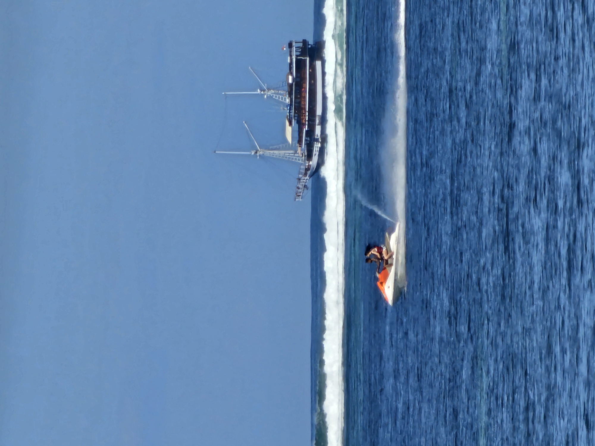 An image depicts a picturesque scene of the ocean with a clear blue sky above. In the foreground, two individuals are riding a jet ski, creating a trail of white spray as they move across the water. In the background, a large, traditional sailing ship with multiple masts and sails is visible, navigating the waves. The ocean exhibits a vibrant blue hue, with white waves crashing in the middle ground, suggesting a coastal environment. The scene is set in the waters off the coast of Denpasar, Indonesia, a popular tourist destination known for its beautiful beaches and water activities.
