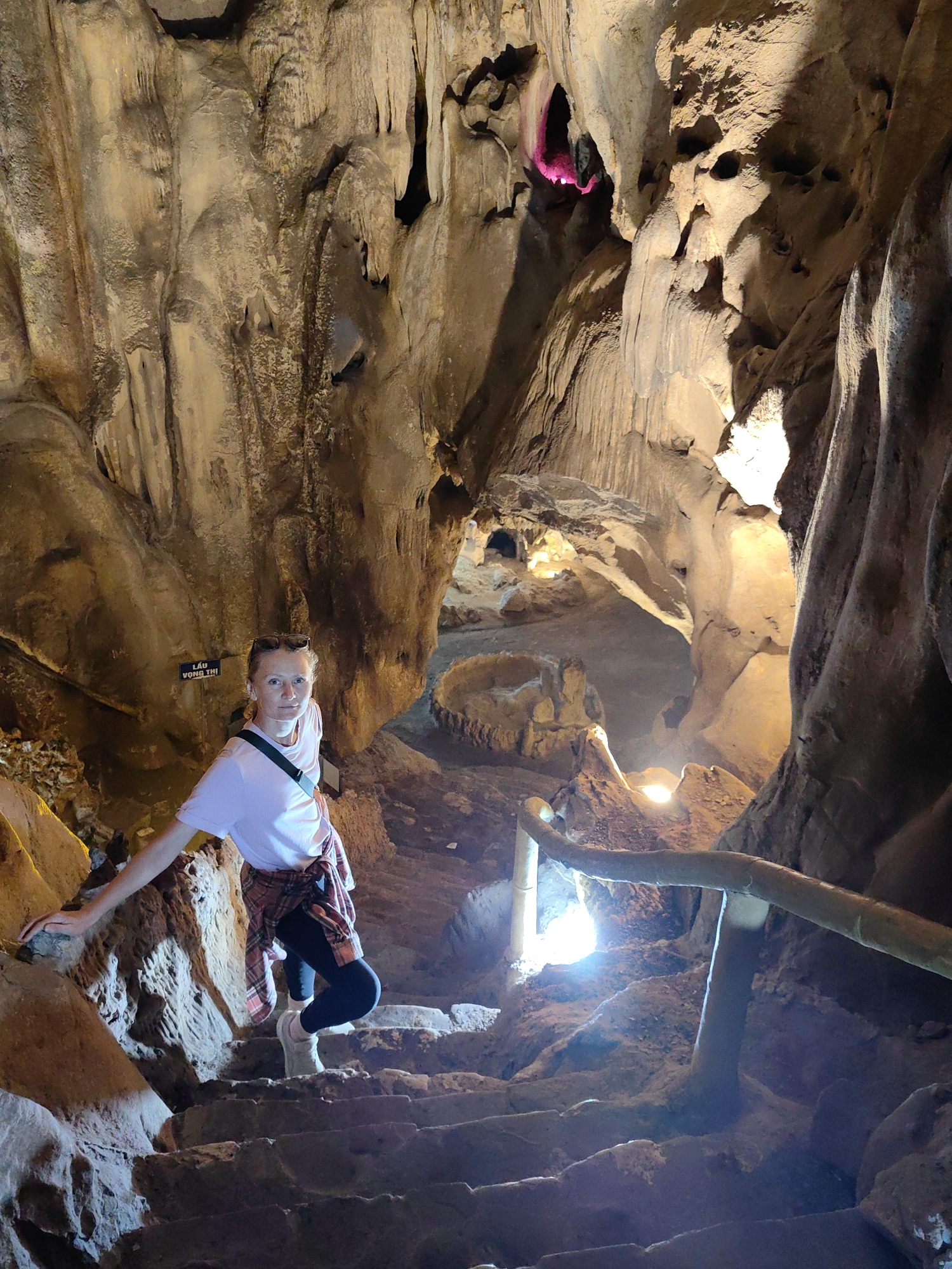 A Caucasian woman is descending stairs within a cave. She wears a white t-shirt, black leggings, a plaid shirt tied around her waist, and light-colored shoes. Her left hand touches the cave wall, while her right hand is partially obscured. The cave's walls display formations, with a sign visible at the top-left, bearing Vietnamese text, suggesting the location may be in Vietnam. A portion of the cave is illuminated with pink lighting. In the middle-right, a wooden handrail is installed along the stairs. The stairwell leads downward and the location is Lạc Giao, Quảng Lạc, Vietnam.