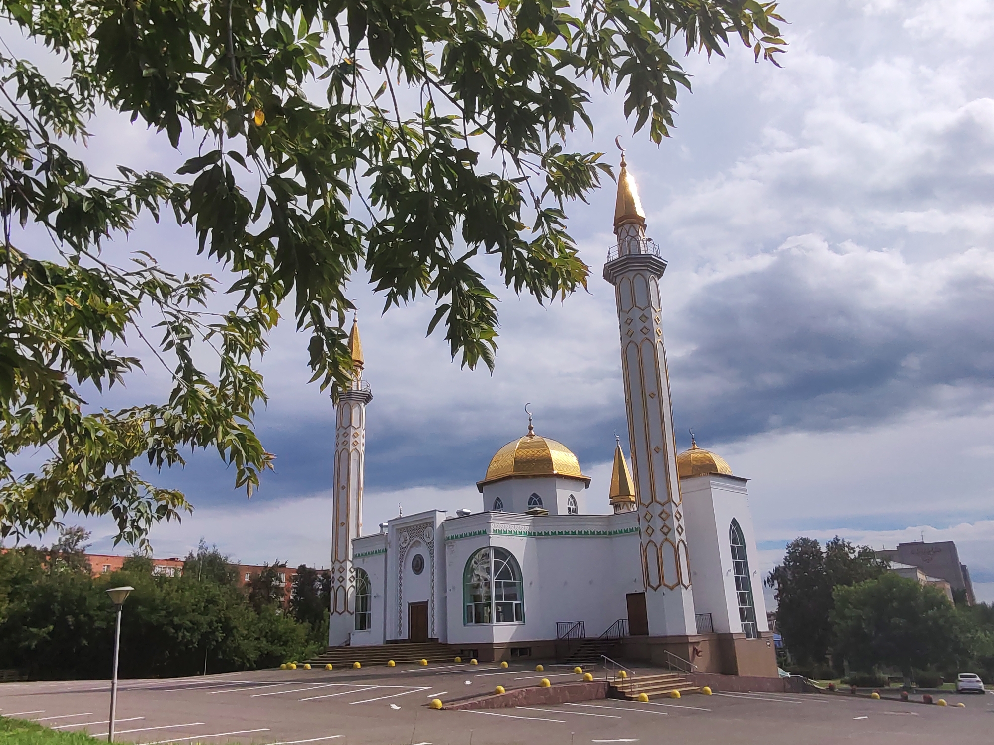 The image shows the "Naim" mosque in Izhevsk, Russia. The mosque is a white building with golden domes and minarets. It is surrounded by trees and a parking lot. The sky is cloudy. A white car is parked in the background. The image is taken from a low angle, looking up at the mosque.