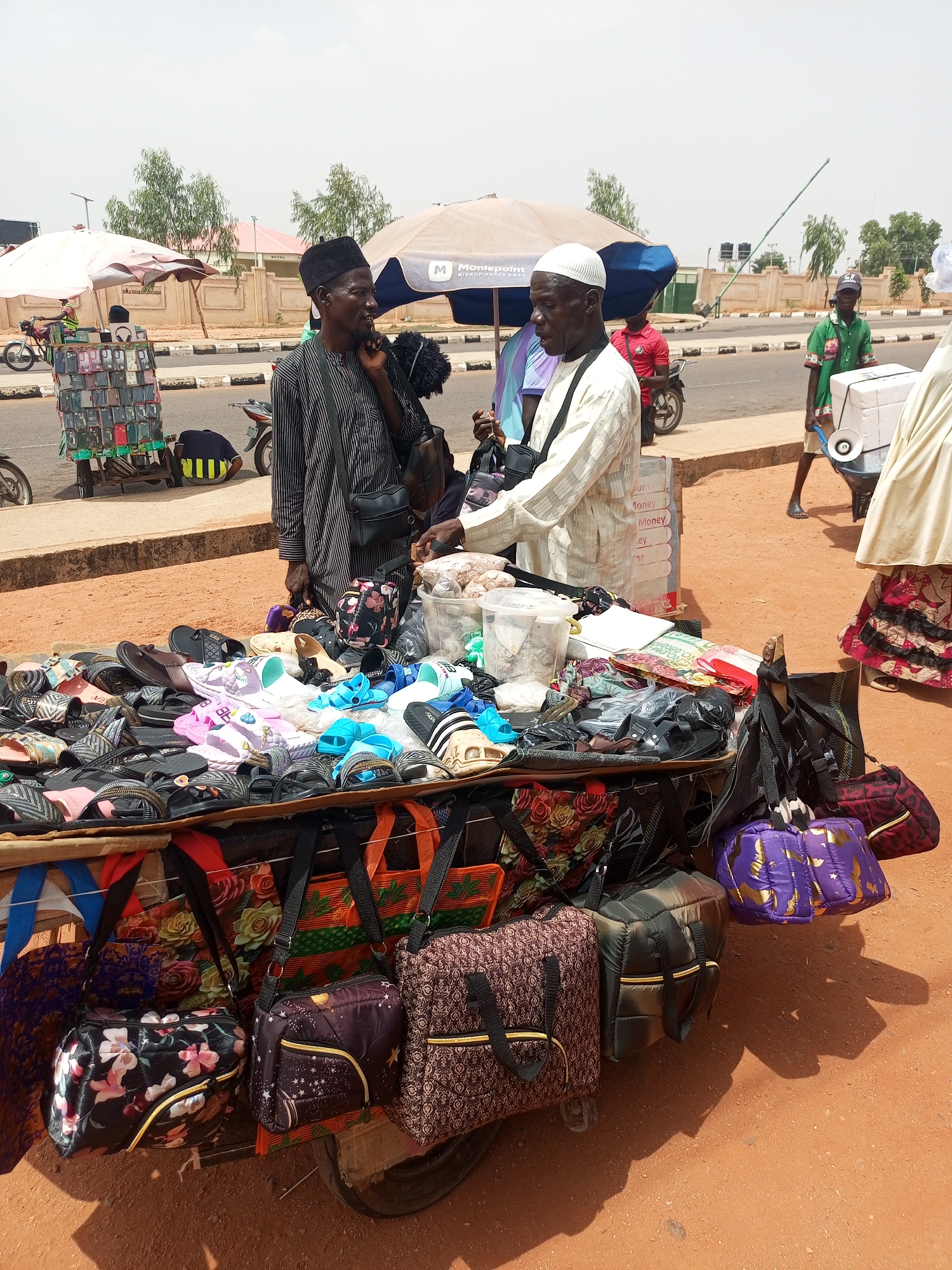 A street scene in Gombe, Nigeria, shows a mobile vendor standing behind a cart piled with sandals, bags, and miscellaneous items. He is a dark-skinned man wearing a white skullcap and a white striped shirt and trousers, interacting with a customer. The customer is a dark-skinned man wearing a black skullcap and a striped shirt with a black bag slung over his shoulder. The cart is heavily laden with a variety of sandals and bags, which hang over the sides.
In the background, other vendors are visible, including one selling phone cases and another using a wheelbarrow. Motorbikes pass on the road. The setting appears to be a commercial area, under a partly cloudy sky. The ground is red earth.