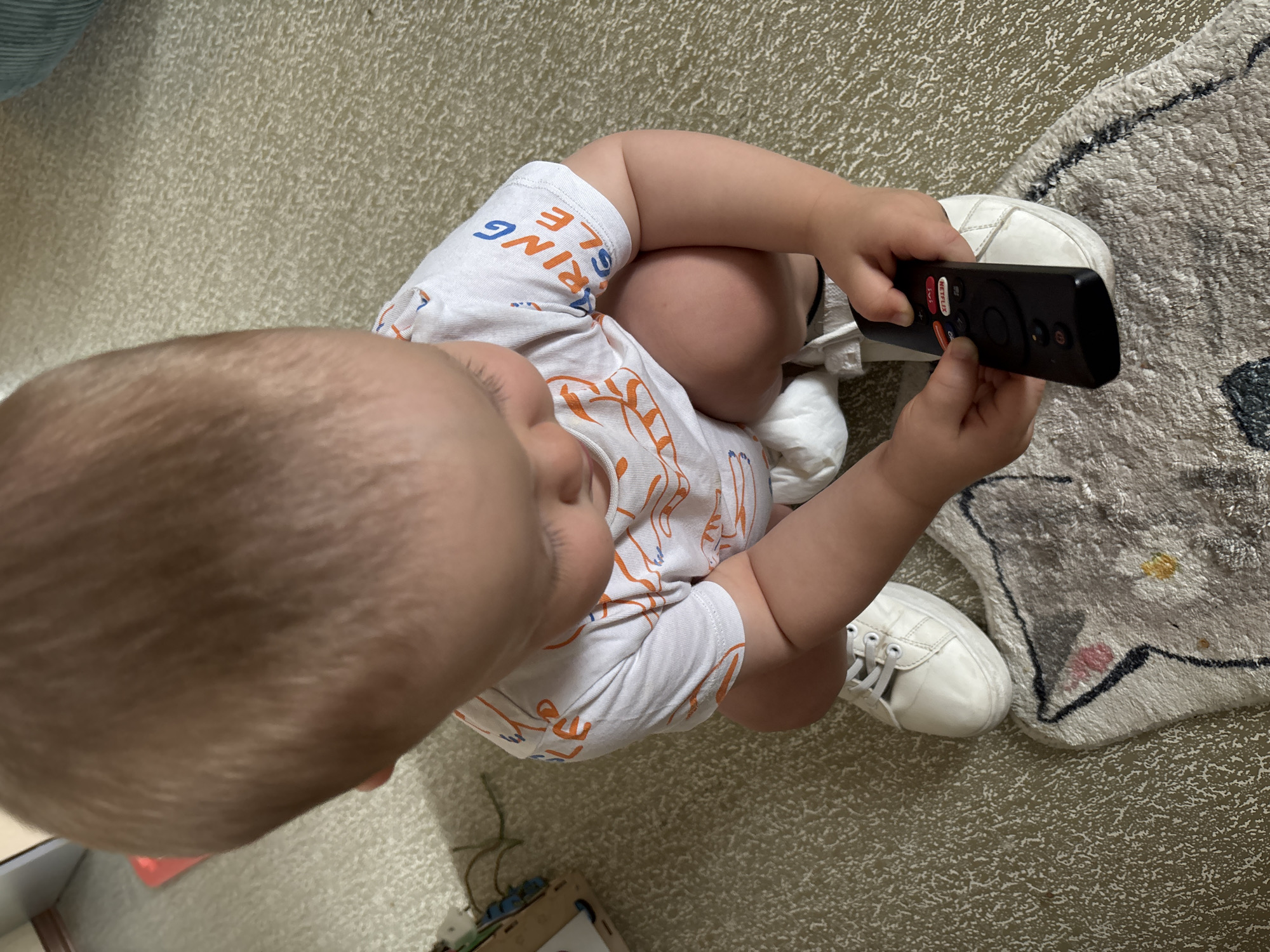 A young child, seen from an overhead perspective, sits on a patterned rug and carpeted floor. The child is wearing a white onesie with an orange graphic print and white sneakers. They are holding a black remote control, appearing to press buttons on it. The remote has a prominent red Netflix button and other circular buttons. The child's head is tilted downwards, and their face is partially obscured. The scene appears to be indoors, with natural light suggesting daytime. There are no obvious indications of weather or specific time of day beyond daytime. The context of Naberezhnye Chelny, Russia is provided but not visually evident in the image itself. The overall impression is one of a child engaged with technology.