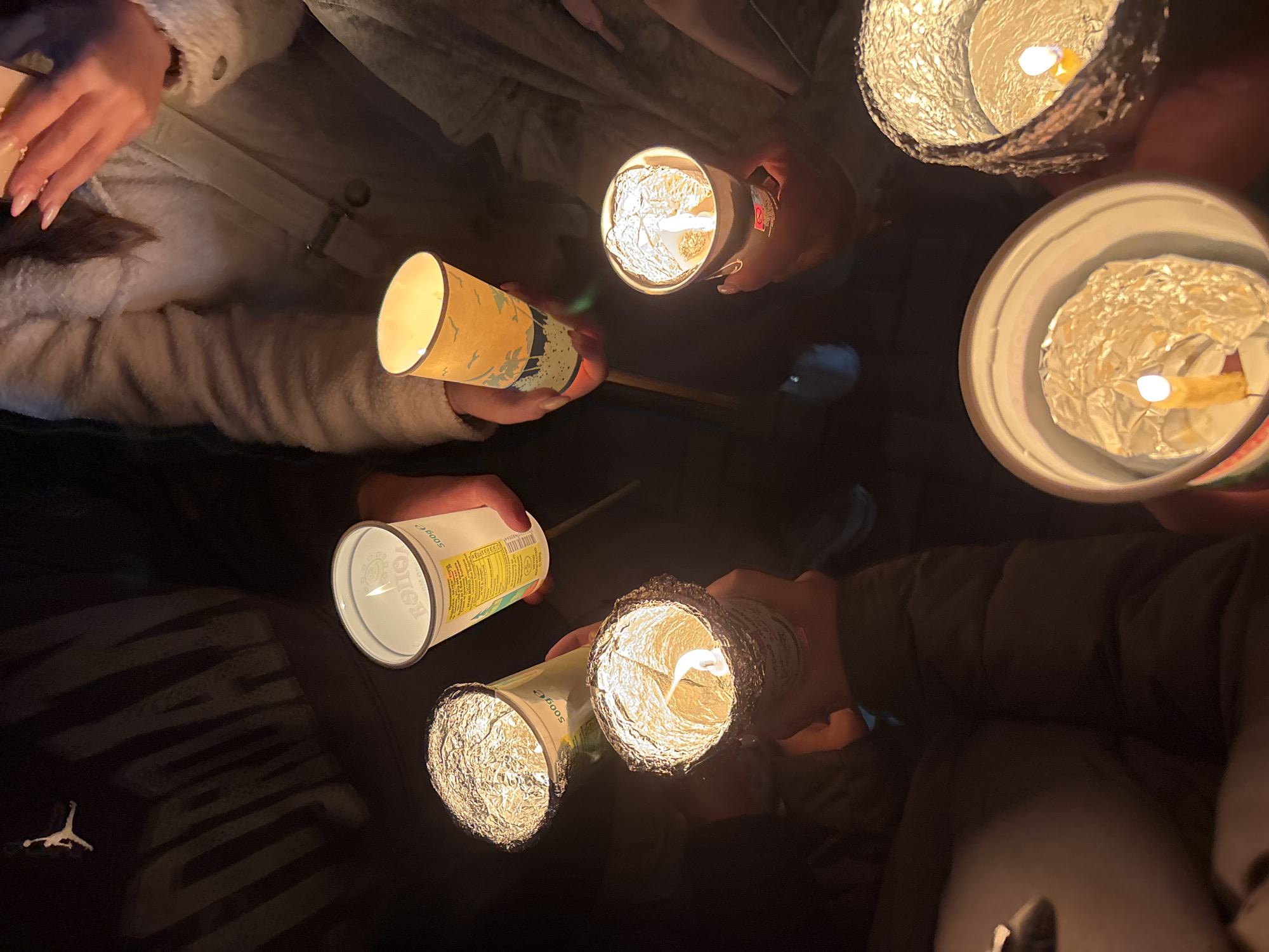 The image shows a group of people holding homemade candles, likely during a vigil or gathering. The candles are in cups, some wrapped in aluminum foil, with lit wicks. The scene appears to be dimly lit, suggesting it is either nighttime or in a darkened indoor space. Individuals are wearing various coats, and a Jordan logo is visible on someone's clothing. Location is unknown.