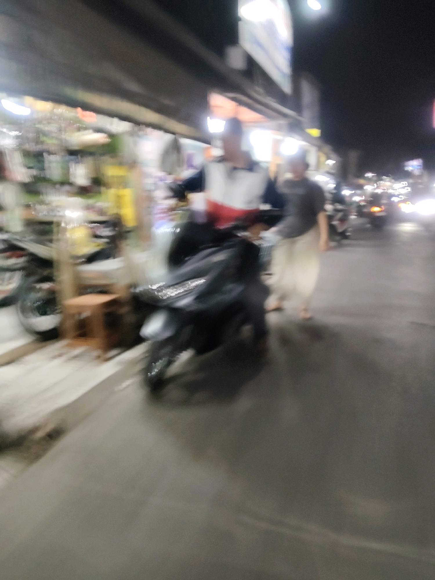 A night scene in Mekarmukti, Indonesia, shows a street with shops on the left. A person in a white, red, and blue shirt is on a dark-colored motorcycle. Another person in a dark shirt and light-colored pants is walking on the right. The street is wet, reflecting the lights of passing vehicles, which are blurred due to motion. The overall image is out of focus, creating a streaky effect.