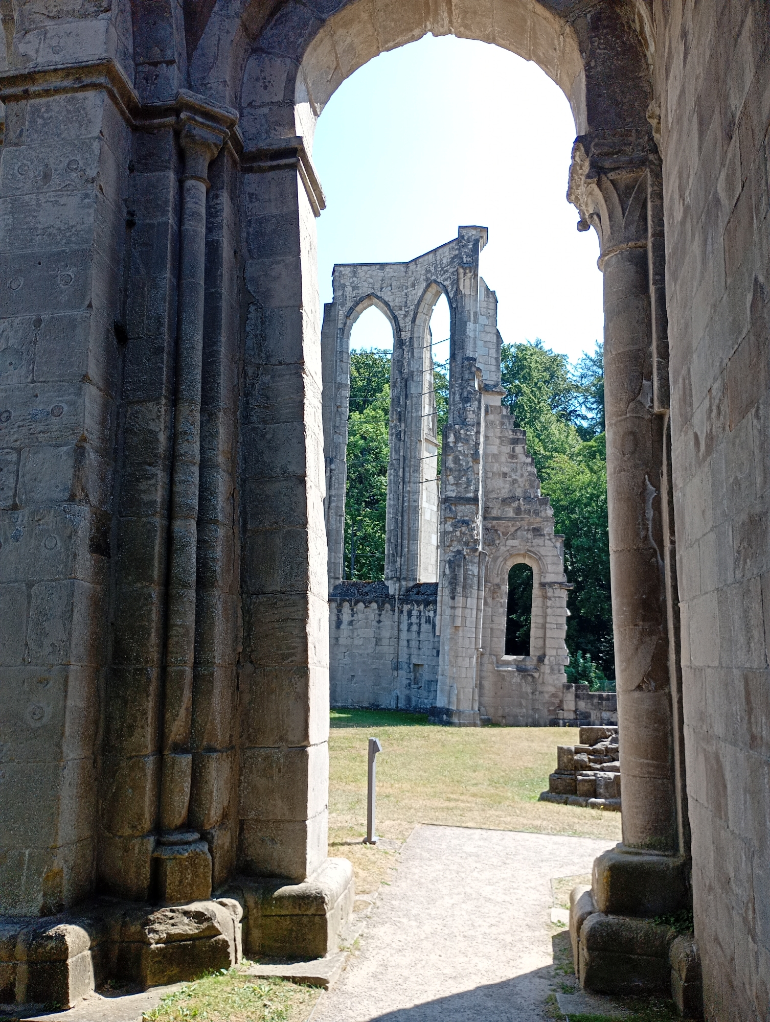 This is a full-shot, eye-level, outdoor photograph of the ruins of a large stone building, likely an abbey or church, with trees and a clear sky in the background. The ruins are seen through an arched entryway formed by massive stone columns. The stone walls are weathered and show signs of decay, with many missing stones and crumbling sections. The building's architectural style appears to be Romanesque or Gothic, with arched windows and supporting structures. The ground in front of the ruins is covered in dry grass and a gravel path. A small, thin metal pole is visible in the grassy area. The scene is bathed in sunlight, casting shadows from the architectural elements onto the ground. The photograph was taken at the Walkenried Abbey in Walkenried, Germany. There are no people or animals visible in the image.