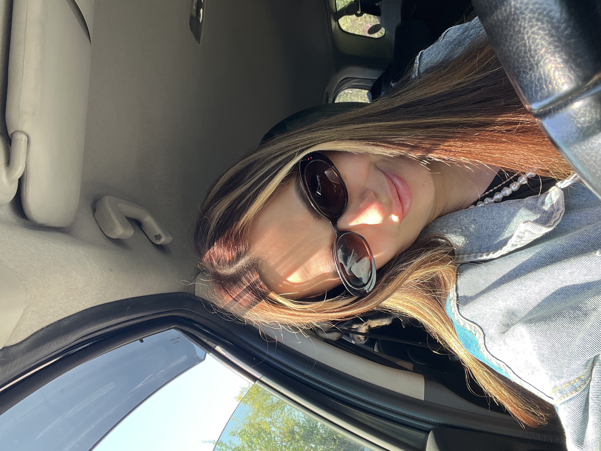 A woman is sitting in the driver's seat of a car, wearing sunglasses and a denim jacket over a black top with a pearl necklace. The car's interior is visible, including the steering wheel, window, and sun visor. Outside the window, there are trees.