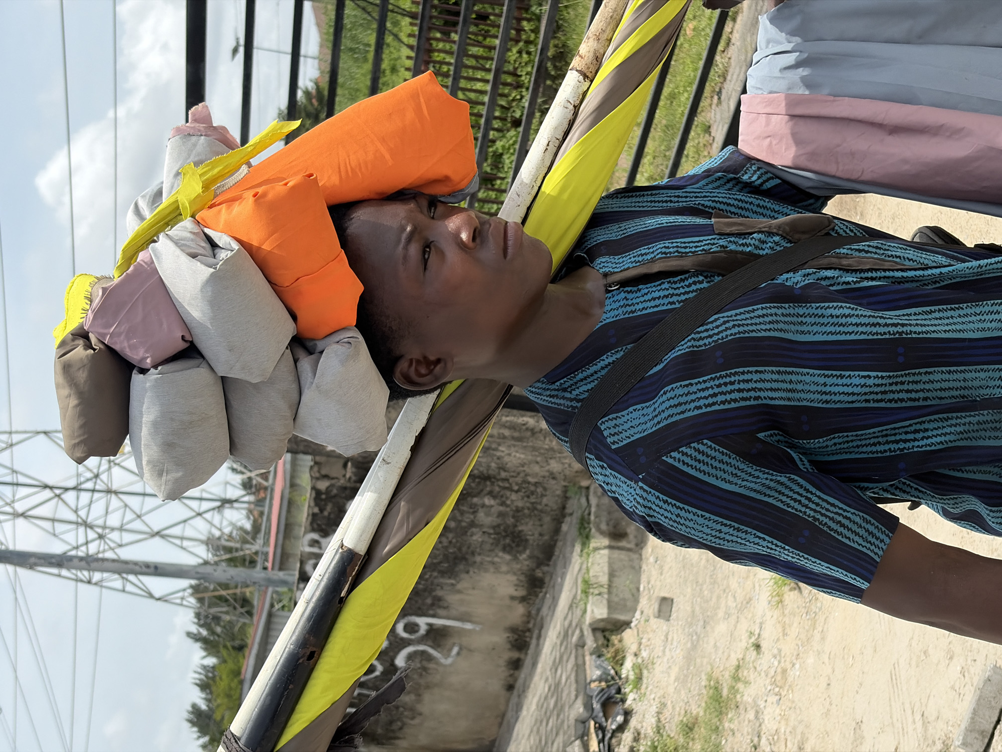 A young man in Igboefon, Nigeria, is carrying a load of rolled fabrics on his head. The fabrics are various colors, including orange, gray, pink, yellow, and brown. He is wearing a striped blue shirt and a strap is across his chest. He is looking upwards. Wooden poles are visible near his head. The background shows a fence, power lines, trees, and a concrete wall.