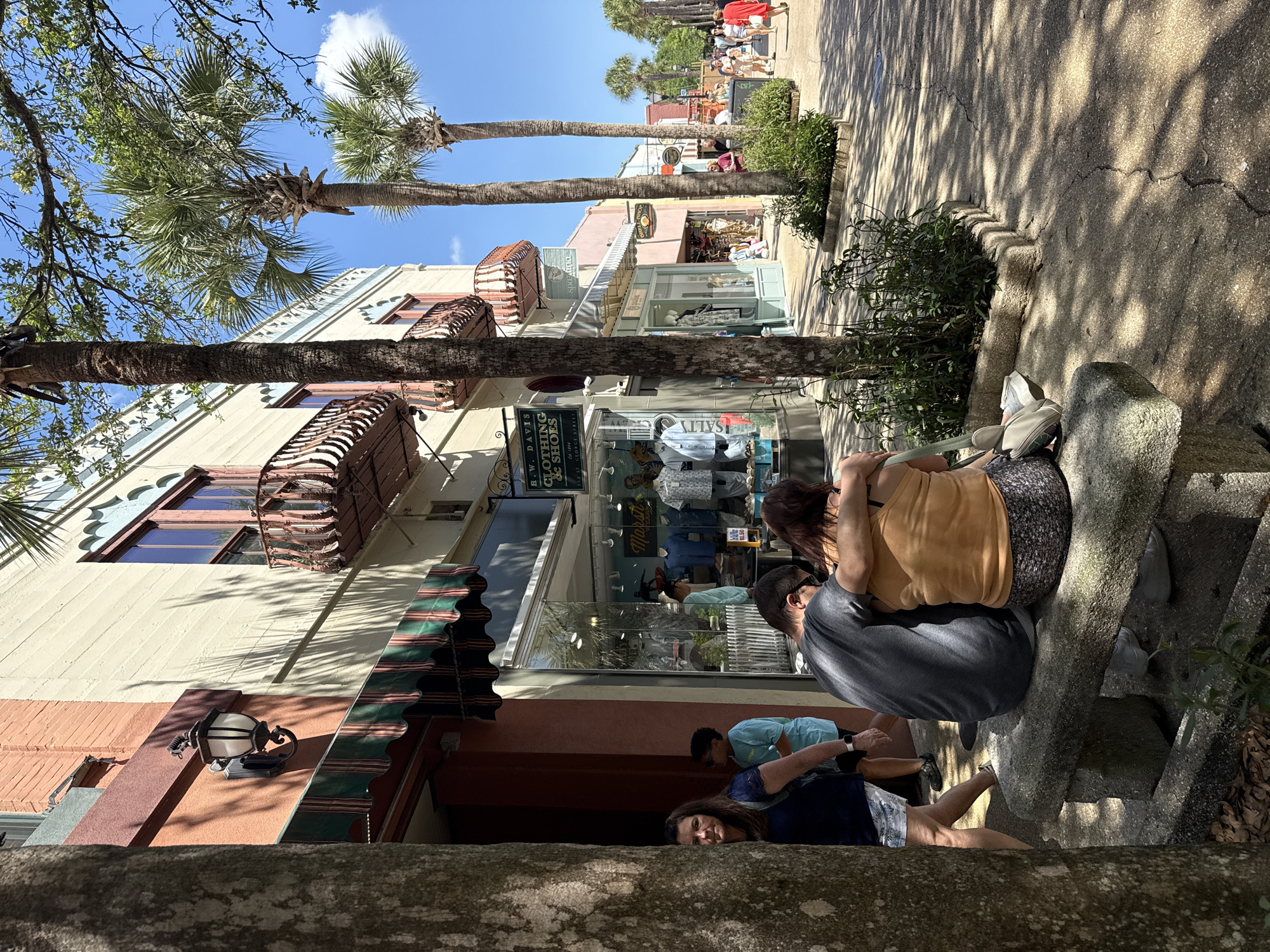 The image depicts a street scene in Saint Augustine, Florida, United States. The view is oriented upwards, focusing on a row of buildings with architectural details such as balconies and awnings. Palm trees line the street, and a group of people are visible in the foreground, seemingly resting or interacting on a stone structure. Shop windows display clothing, and people stroll along the sidewalk. Sunlight casts shadows on the street.