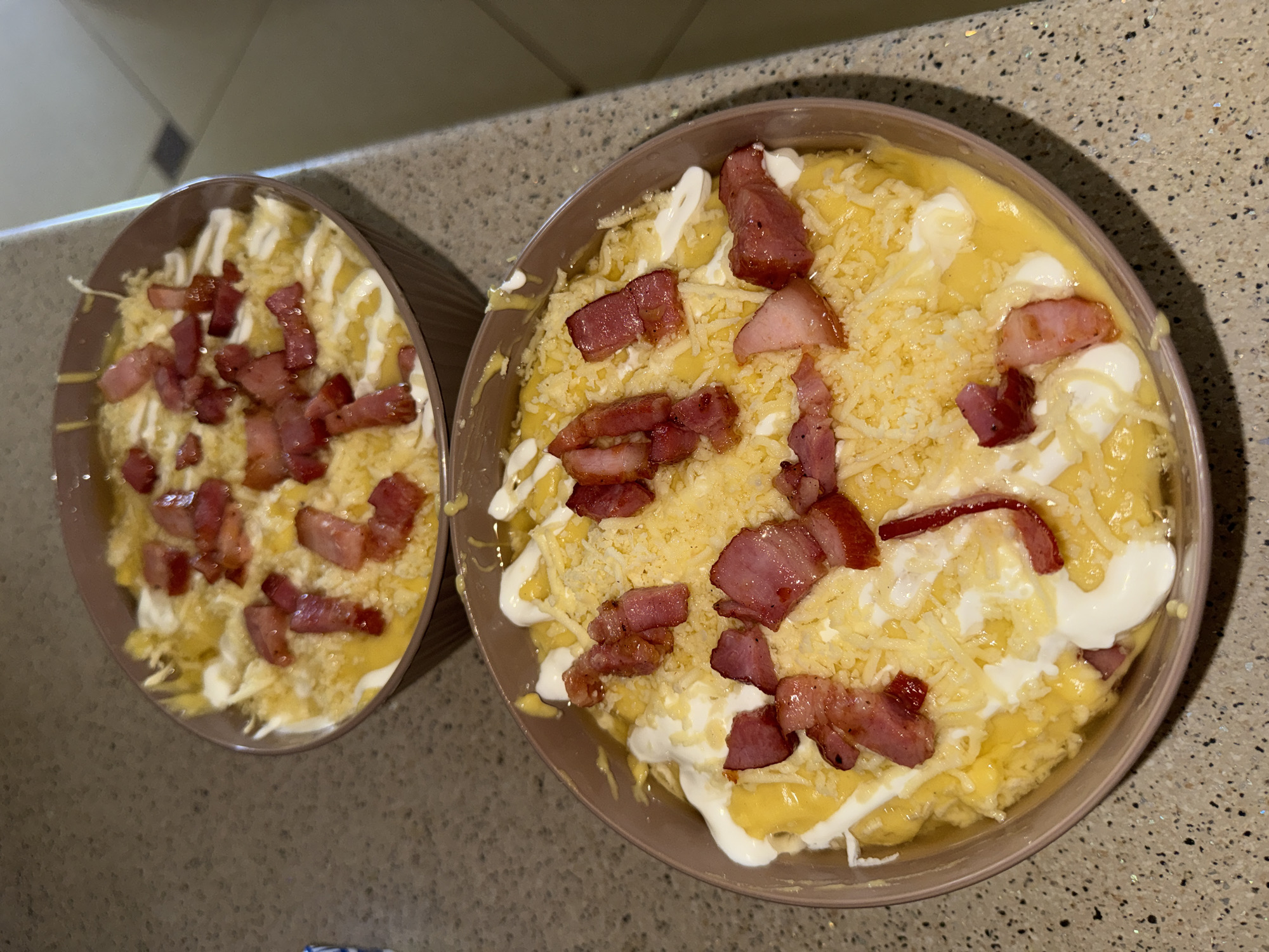 Two bowls of macaroni and cheese topped with shredded cheese, crispy bacon bits, and a drizzle of white sauce are displayed on a speckled countertop in Horbanivka, Ukraine. The bowls are light brown with a ribbed texture. The lighting suggests an indoor setting, possibly a kitchen. No people are visible in the image.