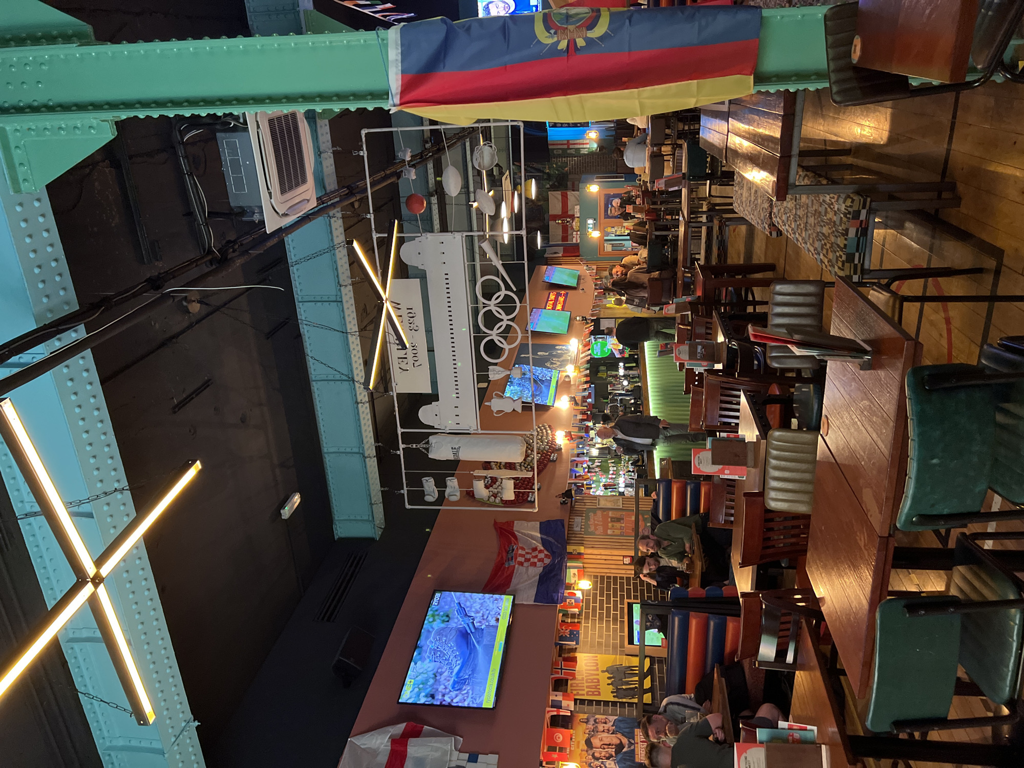 This is an indoor wide shot of a sports bar with wooden floors and tables. A large green structural beam is visible on the right side of the frame, extending from the ceiling to the floor. The ceiling features exposed ductwork and industrial-style lighting. In the background, a long bar with bottles and a television screen is visible. Various flags, including the Croatian flag and the English flag, are displayed on the walls. Several patrons are seated at tables, some eating and drinking, while others are engaged in conversation. A man in a black jacket and blue jeans stands near the bar, talking on his phone. A large flag with vertical stripes of red, yellow, and blue hangs prominently on the right side of the image. In the foreground, several wooden tables and chairs are arranged. The overall atmosphere appears casual and lively, suggesting a sports-watching event or a gathering.
