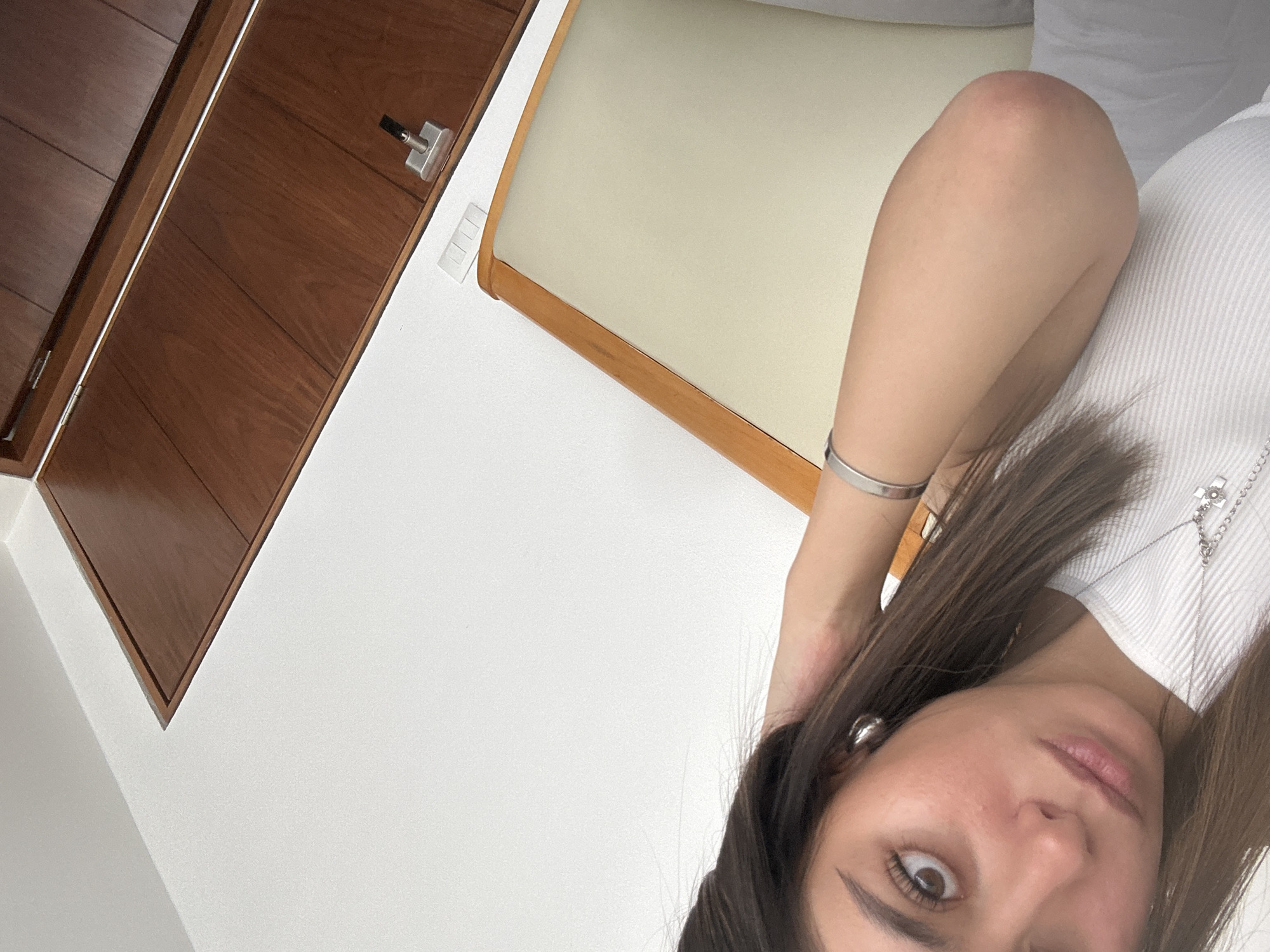 The image captures a close-up, angled view of an adult female in what appears to be an indoor setting. The camera is positioned as if taking a selfie, with the subject's gaze directed towards the lens.
The central subject is a woman with long, dark brown hair that partially covers her left eye and shoulder. She is wearing a white ribbed top or sweater. Her accessories include a silver bangle bracelet on her right wrist, silver hoop earrings, and a silver necklace with a visible cross pendant resting on her chest. Her right eye is wide open, while her left eye is mostly obscured. Her lips are slightly parted, revealing a natural pink hue, and her eyebrows are dark and well-defined.
In the background, to the upper left, a wooden cabinet or door with horizontal paneling and a modern silver handle is visible, slightly ajar. Next to it, part of a piece of furniture, likely a chair or bench, with a light-colored (cream or off-white) upholstered cushion and a wooden frame, can be seen. A small, white, rectangular object with several vertical ridges is affixed to the wall between the cabinet and the cushion. The rest of the background consists of plain white walls or ceiling, suggesting a brightly lit interior. The overall image has a clean and minimalist aesthetic. There is no visible text.