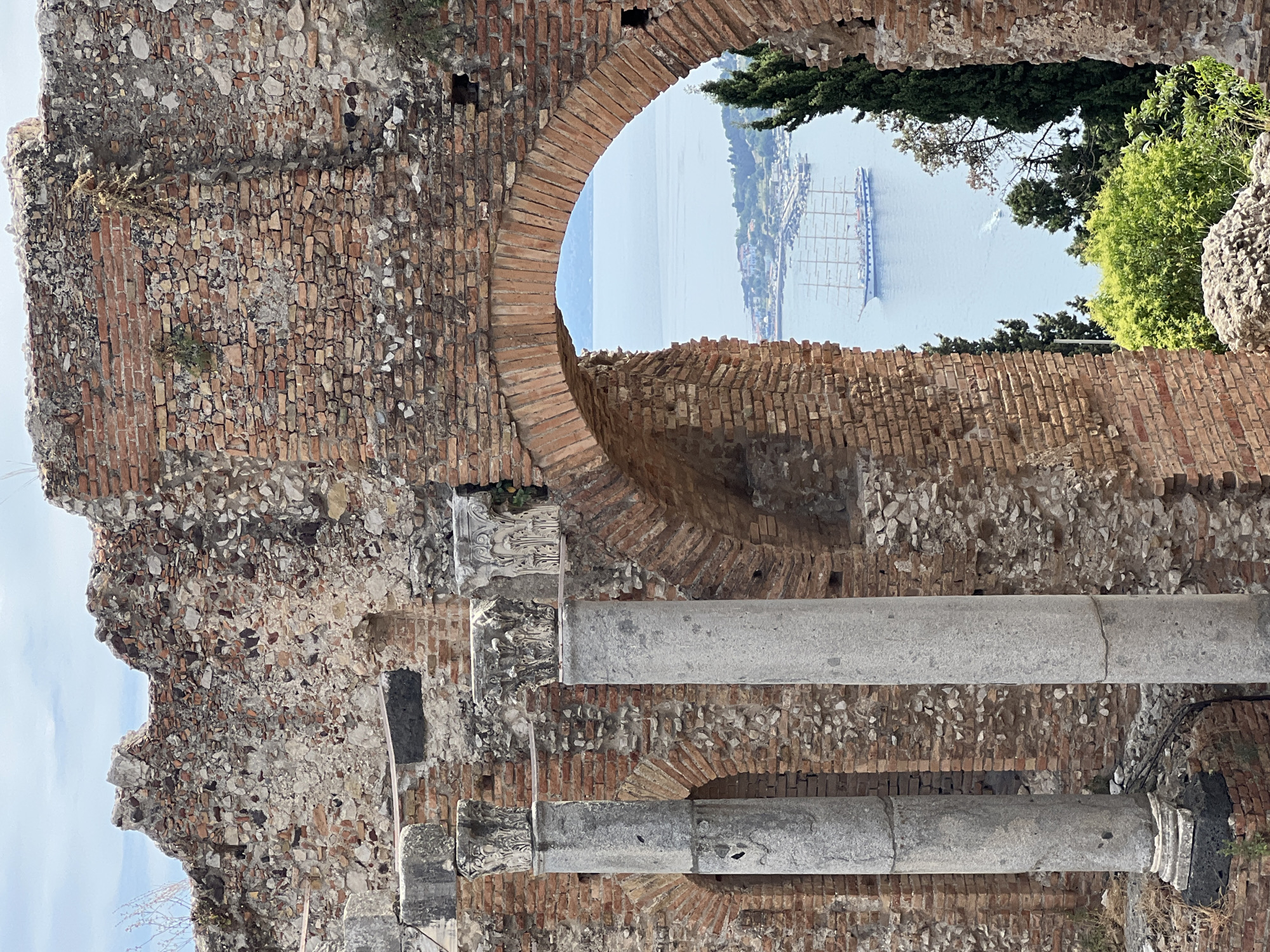 This is an image of the ancient Roman Theatre of Taormina, Italy. The photo is taken from within the ruins, looking out through an arched opening. The foreground is dominated by weathered, ancient brickwork and large stone columns. Some of the brickwork shows signs of vegetation growing amongst the stones, suggesting age and exposure to the elements.
Through the archway, a bright, clear body of water, likely the Ionian Sea, is visible. On the water, a large, multi-masted sailing ship is anchored. Beyond the ship, the coastline is lined with buildings and greenery, indicating a populated area. The sky above is a pale blue. The time of day appears to be daytime, likely morning or afternoon, given the bright light and lack of shadows. The weather seems fair and sunny.
The overall impression is one of historical grandeur combined with a beautiful natural landscape. The scene evokes a sense of timelessness, contrasting the ancient man-made structures with the enduring beauty of the sea and coastline. There is no visible text in the image.