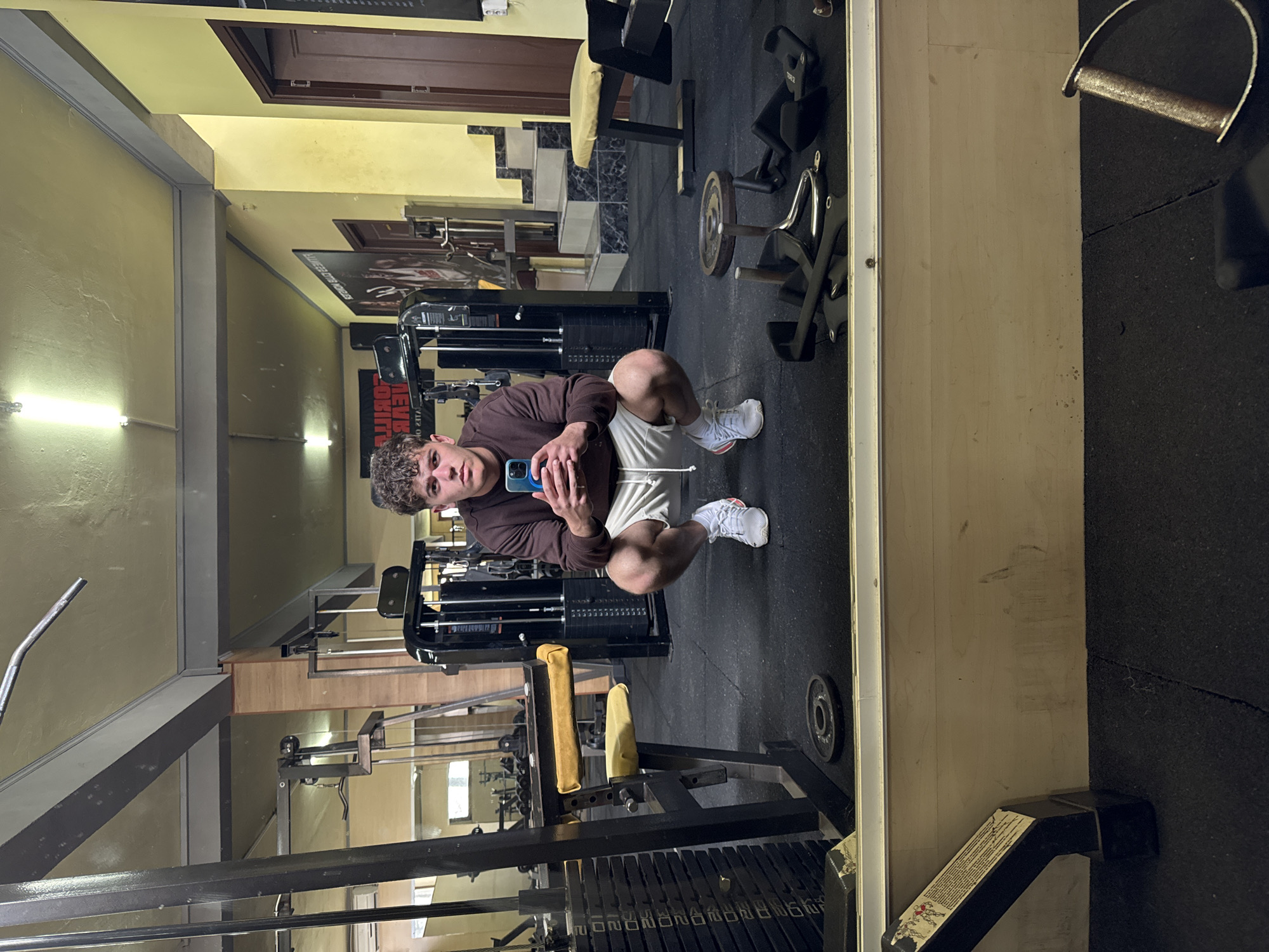 A man squats in front of a mirror in a gym in Goce Delcev, Bulgaria. He wears a brown sweatshirt, white shorts, and white sneakers, and holds a blue phone in his left hand. Various weight machines and free weights are visible in the background. The gym has black rubber flooring and light yellow walls.