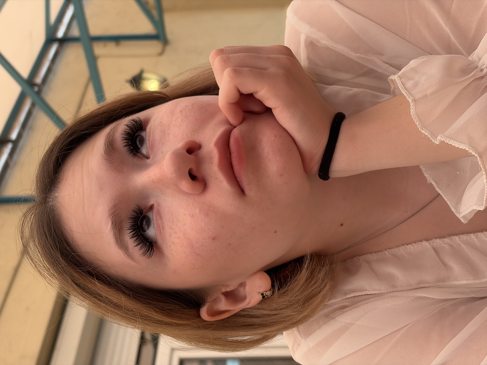 A young Caucasian female with blonde hair and light skin with freckles is pictured in a close-up shot, looking upwards and to the right. She has on full, dramatic false eyelashes and is resting her chin on her right hand, which is supported by her left arm. She is wearing a sheer, light pink blouse with ruffled details at the neckline and cuffs. A thin silver necklace is visible around her neck, and she has a small, dark stud earring in her left ear. The background is blurred but appears to be an outdoor seating area with a light-colored awning supported by green metal beams, and a lantern is visible in the background. The lighting suggests it is daytime.