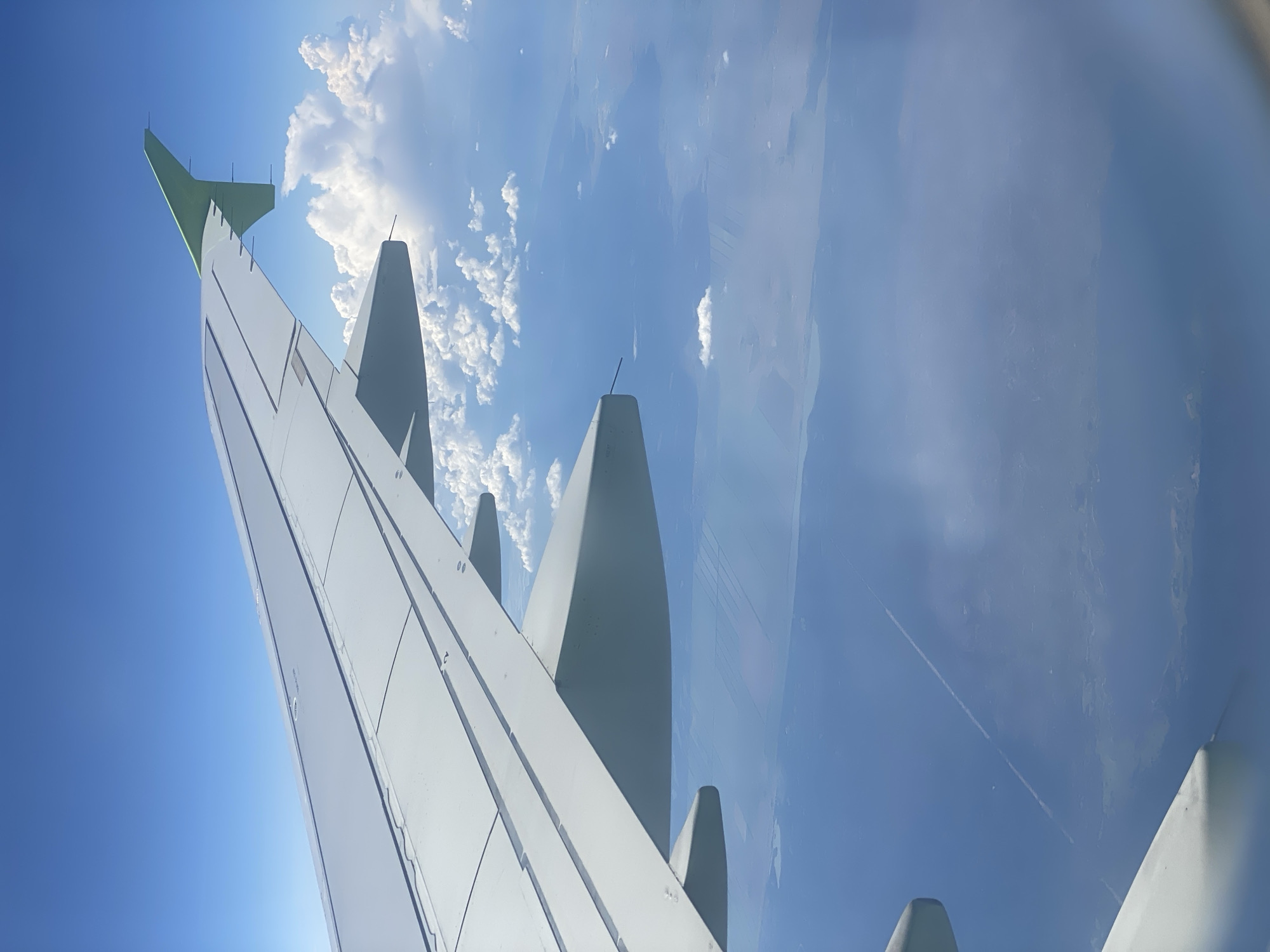 An aerial view from an airplane window shows a white airplane wing extending across the frame. Below the wing, a vast expanse of blue water stretches to the horizon, dotted with clouds. The water surface displays subtle patterns, possibly indicating land formations or cultivated fields beneath the surface. In the distance, a faint white contrail is visible, extending across the sky. The overall scene depicts a serene flight over water under a clear blue sky with scattered clouds. The location is not specified as Kaip, Russia.
