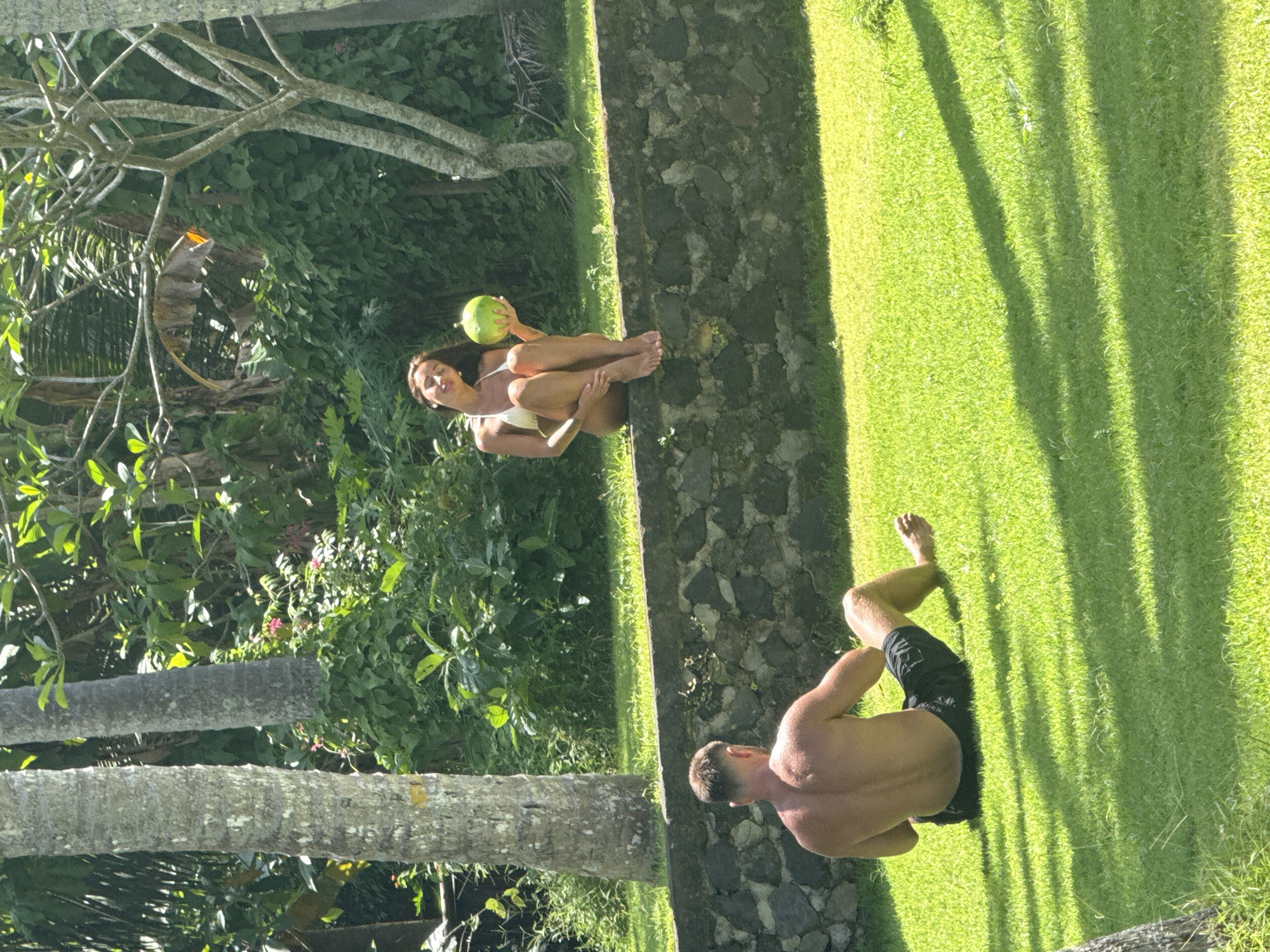 A woman in a white bikini sits on a stone wall holding a green fruit. A man without a shirt is lying on the adjacent grass. The location is likely near Karangasem, Indonesia given the lush tropical plants and palms visible in the background.