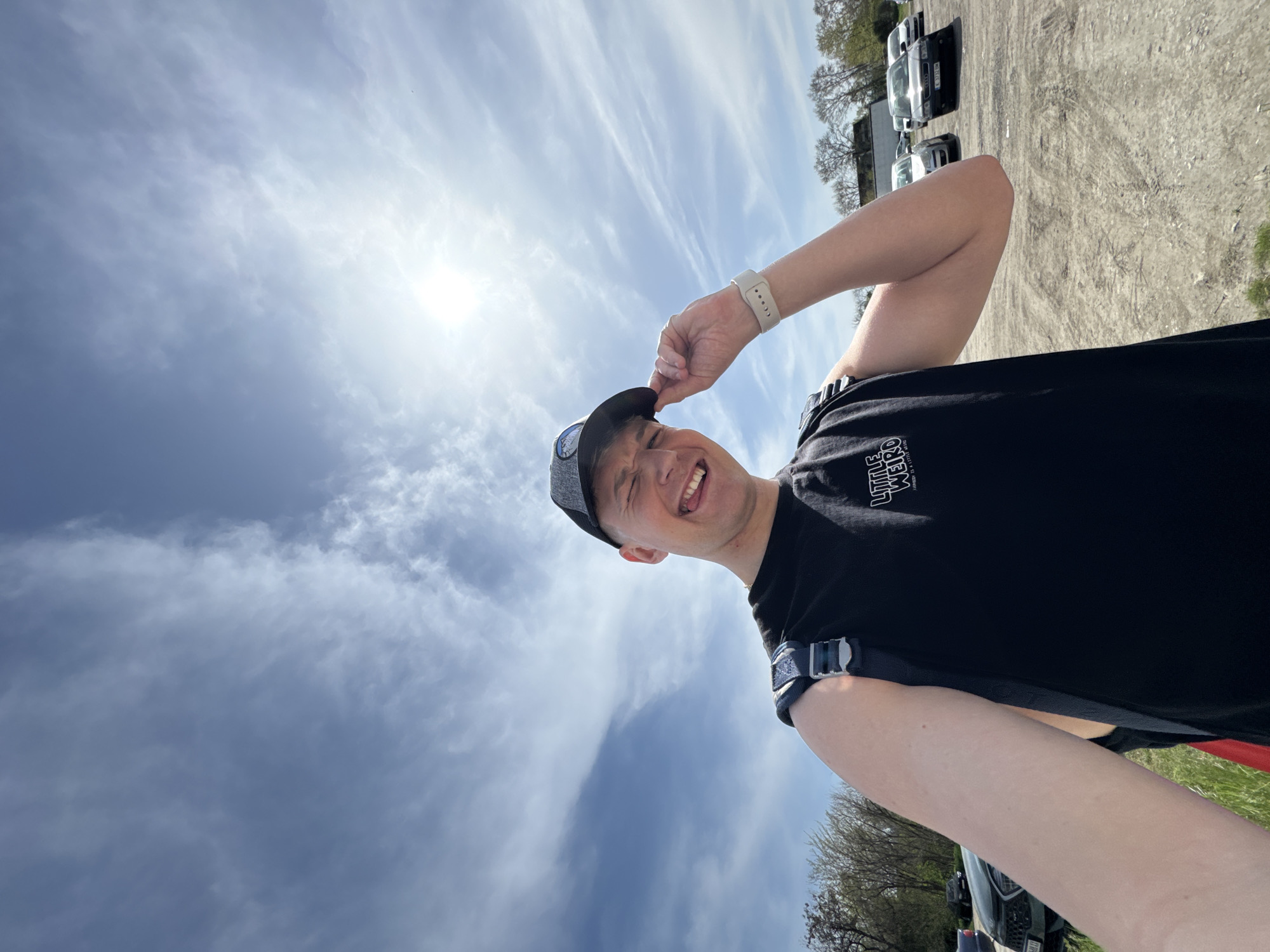 This media file captures a cheerful young man in an outdoor setting, likely in Plovdiv, Bulgaria, under a bright, partly cloudy sky. The image is taken from a low angle, looking up, and is rotated clockwise, making the horizon appear vertical.
The main subject is a smiling man, likely Caucasian, seen from the chest up. He is wearing a grey baseball cap, which he playfully pulls down with his left hand, and a dark t-shirt. On his t-shirt, the white text "LITTLE WEIRD" is partially visible. He also wears a light-colored smartwatch on his left wrist and a dark strap, possibly from a backpack or harness, is visible over his shoulders. His expression is one of clear joy, with a broad smile, open mouth, and slightly protruding tongue.
The setting includes a vast expanse of blue sky with scattered cirrus and cumulus clouds, indicating good weather. The sun shines brightly from the upper left (in the rotated view), suggesting it is daytime, possibly mid-morning or afternoon. In the background, to the right and bottom of the rotated image, there's a dirt or gravel parking lot where several vehicles, including cars and possibly vans, are parked. Trees with minimal foliage are visible in the distance, suggesting early spring, late autumn, or a deciduous species not yet in full leaf. No specific event is depicted other than the man posing for the camera in a lighthearted manner.