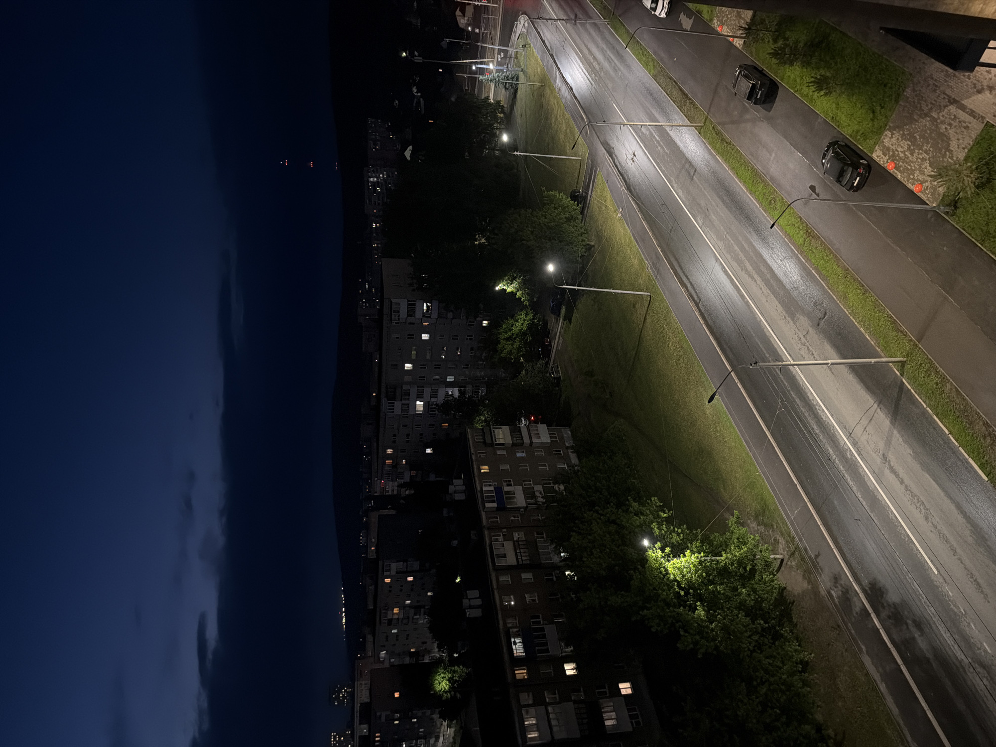 The image shows a nighttime cityscape. The sky is a dark blue with some clouds. A road runs through the center of the image, illuminated by streetlights. There are cars parked along the side of the road and some driving on it. On the left side of the road are buildings with lit windows. There are trees lining the street. The location is Unknown, Unknown.