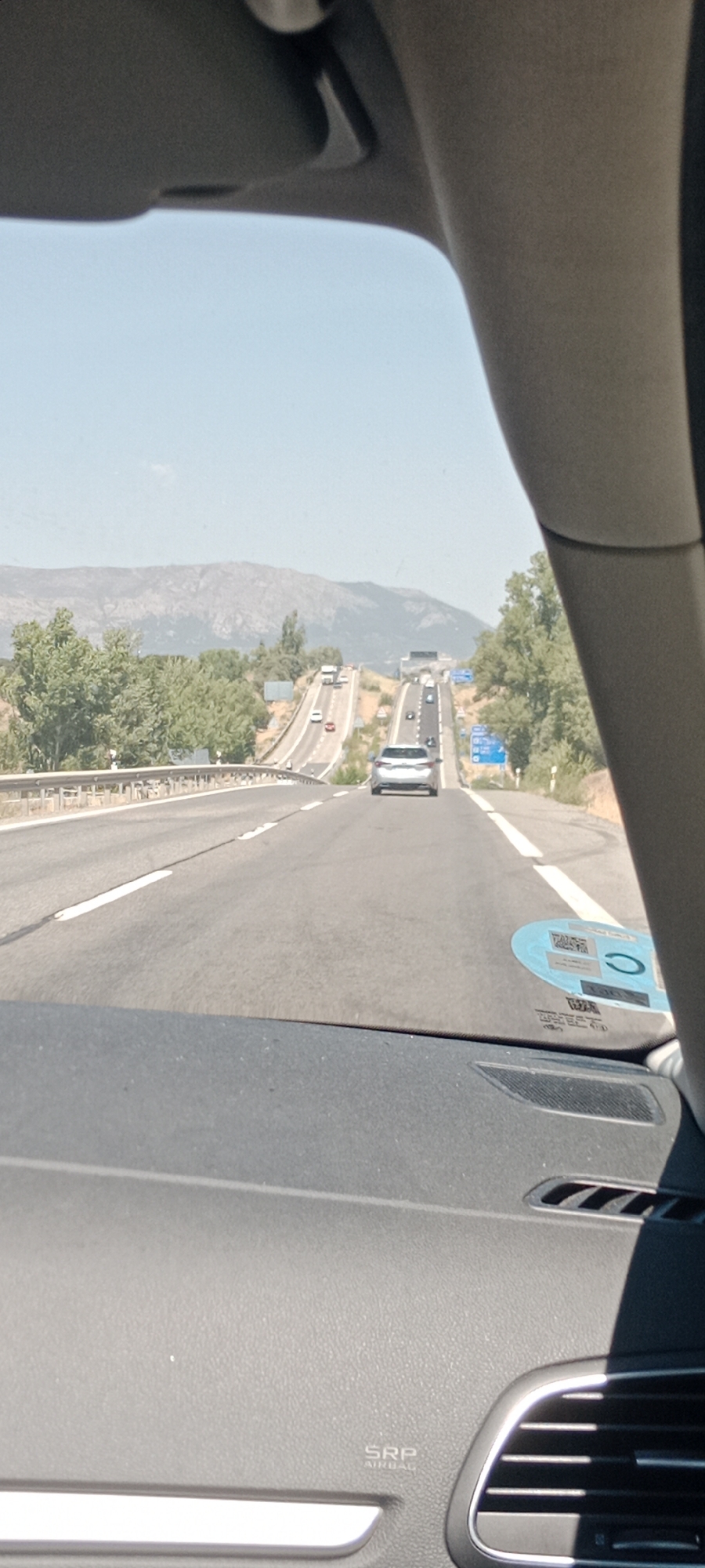 The image shows a view from inside a car driving on a highway. The highway has multiple lanes and appears to be an uphill gradient with a significant incline. There are several cars visible on the highway, traveling in the same direction. The landscape on either side of the highway consists of rolling hills covered in green trees and vegetation, with mountains visible in the distance under a clear blue sky. The interior of the car, including part of the dashboard and windshield, is visible in the foreground. A sticker is affixed to the windshield. This scene is likely located on a highway near Pedrezuela, Spain, given the characteristic landscape and the presence of highway infrastructure in a mountainous region.