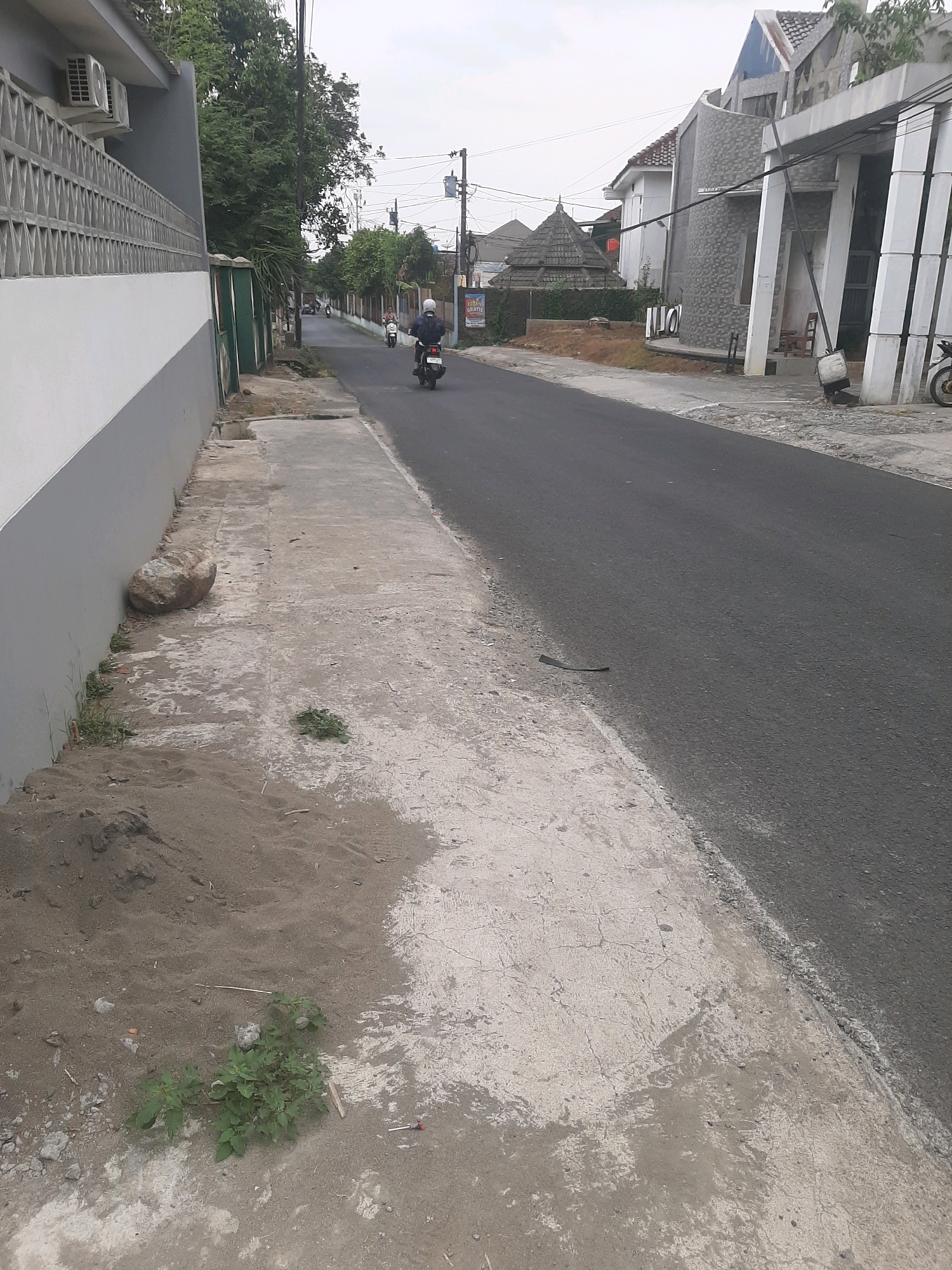 The image depicts a street scene in Sumampir, Indonesia. On the left side of the road is a sidewalk made of concrete, with a pile of sand and a large rock near a wall. A person on a motorcycle is riding down the asphalt road. The road is lined with houses and trees. In the background, there are power lines and a cloudy sky. The overall impression is of a quiet residential street.