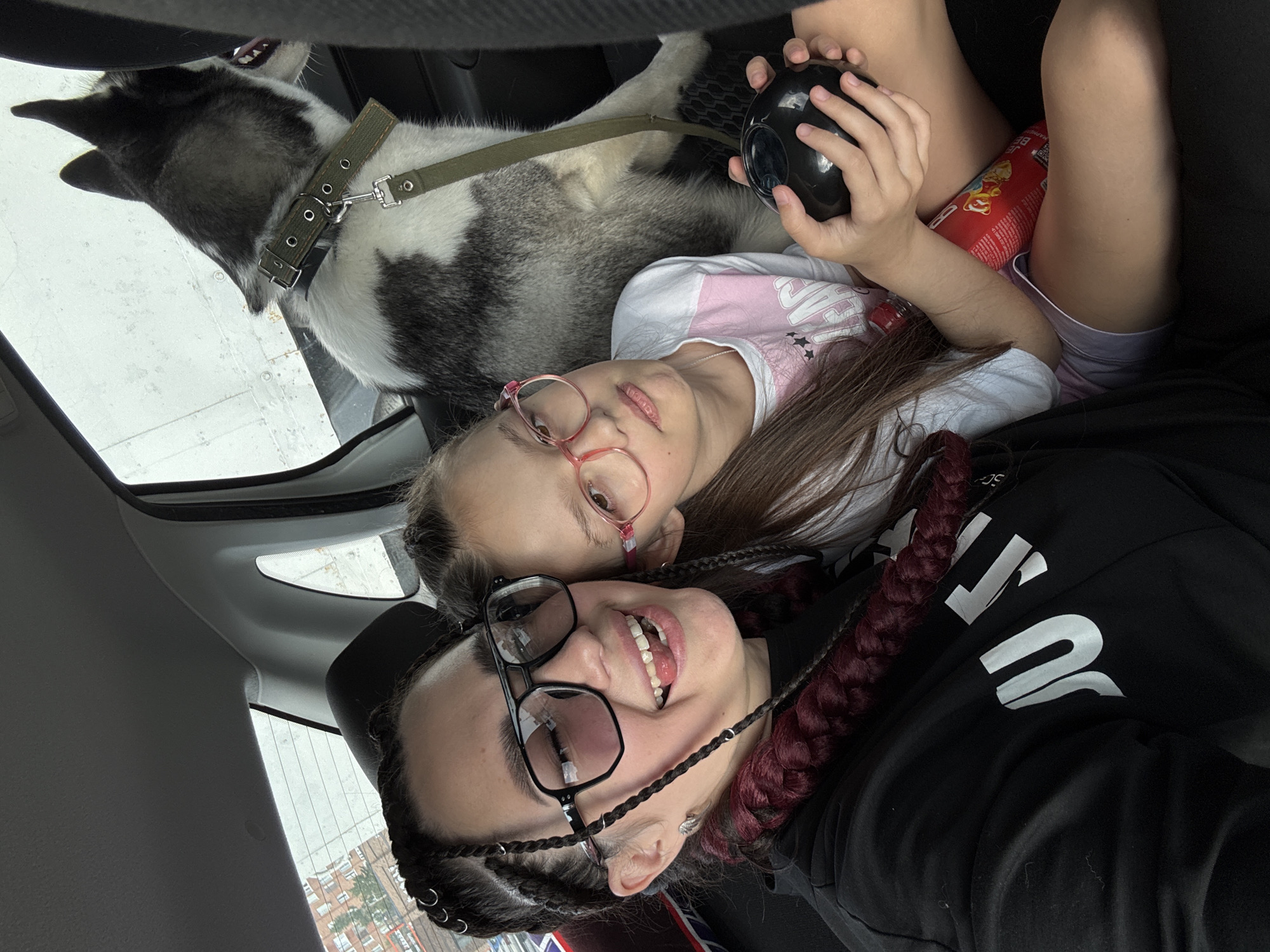 The image shows two young girls and a dog in the back seat of a car. The girl on the left is wearing a black t-shirt, large black-rimmed glasses, and has dark braided hair with red accents and metallic rings. She is sticking her tongue out and smiling. The girl on the right is wearing a pink and white t-shirt and pink-rimmed glasses. She is holding a black ball in her hands and has a bottle of soda next to her. The dog, a husky with black and white fur, is looking out the window. The interior of the car is visible, with the car's headrest and the window showing an urban background with buildings.