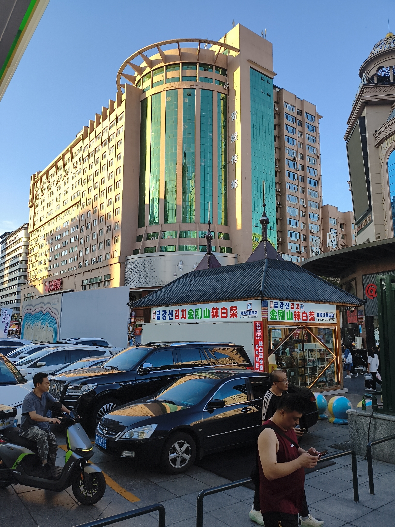 This is a street-level, eye-level, horizontal, medium shot photograph taken outdoors in Mudanjiang, China, likely during the daytime given the bright sunlight. The scene captures a bustling urban environment dominated by a tall, modern building with a distinctive circular, tiered roof structure. The building's facade is a blend of beige-colored concrete and vertical sections of reflective, teal-colored glass, with numerous windows and visible air conditioning units.
In the foreground, several vehicles are parked, including a black SUV, a black sedan, and white cars. A man is seated on a grey and green electric scooter, looking to his left. Two other men are standing on the sidewalk near the parked cars; one is wearing glasses and a black and white striped shirt, and the other is wearing a maroon sleeveless shirt and looking at his phone.
In the middle ground, a small, traditional-looking structure with a dark, tiled, sloping roof and decorative spires sits in front of the large building. This structure appears to be a kiosk or a small shop, with signage featuring Chinese characters and images. The signage includes text such as "금강산 김치" (Geumgangsan Kimchi) and "牡丹江直营店" (Mudanjiang Direct Store), indicating a Korean food vendor. Behind the kiosk, a larger building with more windows and a golden dome is partially visible.
The environment includes a clear blue sky, a paved street or parking area, and a sidewalk. There are also decorative elements on the sidewalk, such as large, colorful inflatable spheres. The overall impression is one of a commercial area with a mix of modern and traditional architectural styles.
