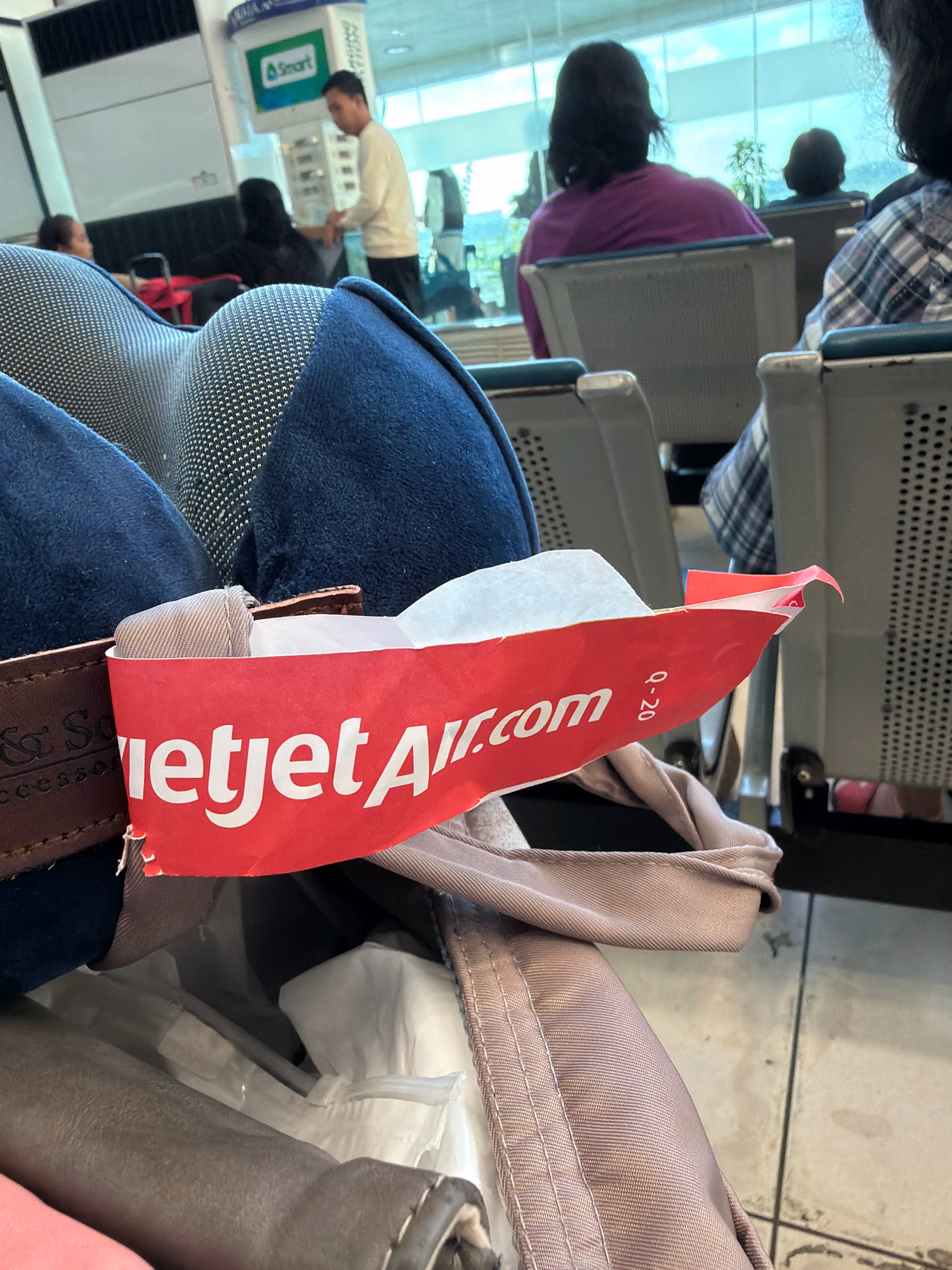The image shows a close-up view of a red tag with white lettering that reads "Jetstar.com" attached to a luggage strap. The tag also has the text "q-20" printed on it. In the background, there are blurred figures of people sitting in an airport waiting area. One person in the foreground is wearing a purple shirt, and another is wearing a plaid shirt. There are rows of seats, and a large window in the background shows a glimpse of the sky. A sign with the word "Smart" is visible on a display in the background, suggesting a location within the Philippines. Considering the presence of Jetstar and the environment, this scene is likely at the Ninoy Aquino International Airport (NAIA) Terminal 1 or Terminal 3 in Pasay City, Philippines, which serves international and domestic flights, including those operated by Jetstar.