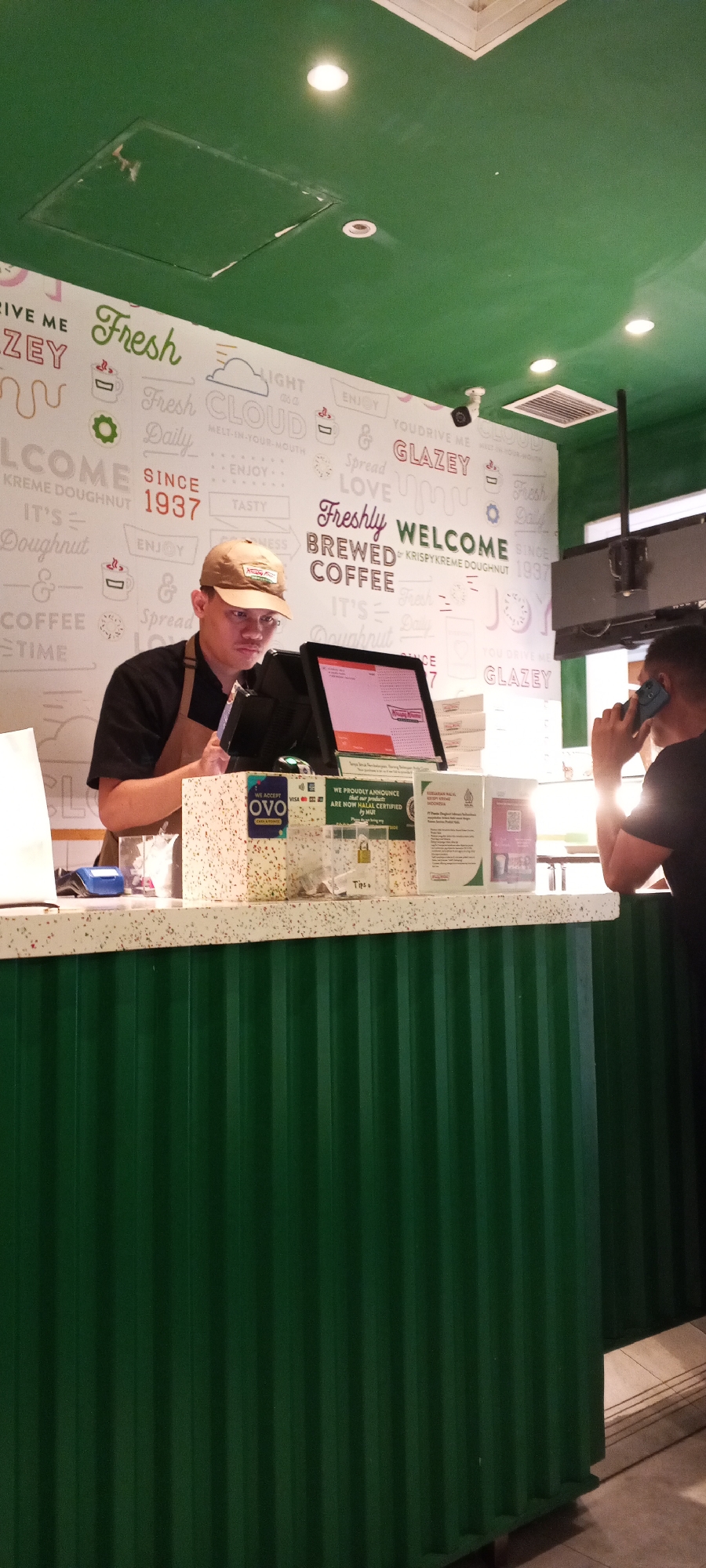 The image shows a Krispy Kreme doughnut store interior in Depok, Indonesia. A male employee wearing a brown apron and a Krispy Kreme branded tan baseball cap is standing behind a counter, looking at a tablet screen. The counter is decorated with a speckled white and grey pattern. In front of the employee, there is a cash register, a card reader, and a tip jar. A box of doughnuts is visible on the counter. To the right of the employee, another man, partially visible from the back, is on the phone. The background features a wall with the Krispy Kreme logo and branding, including phrases like "Freshly," "Brewed Coffee," and "Since 1937." The ceiling is painted green and has recessed lighting fixtures. The counter itself is a vibrant green corrugated metal. Signs on the counter indicate accepted payment methods like OVO and Visa, and a halal certification notice.