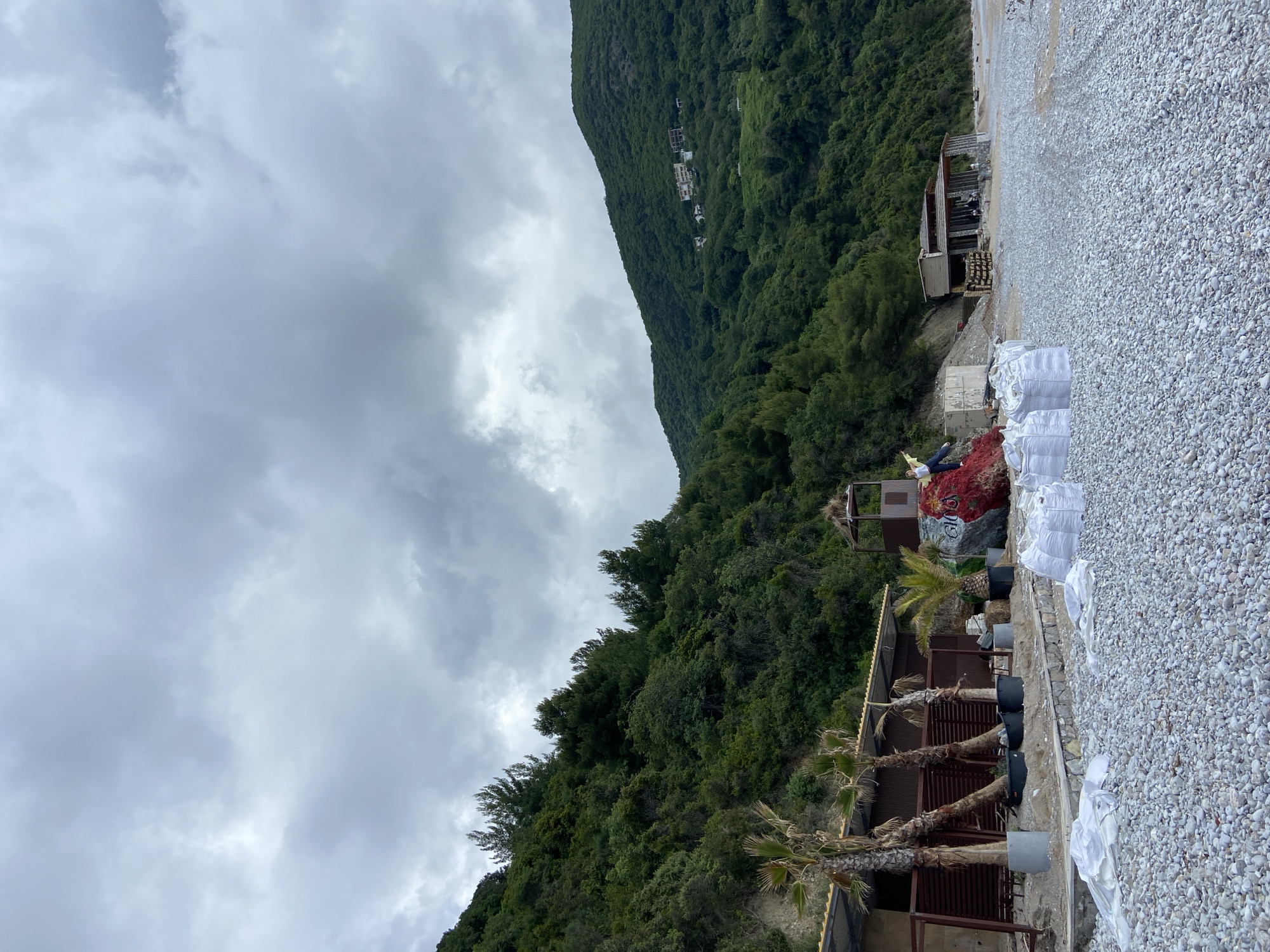The image depicts a scene in Prijevor, Montenegro, featuring a steep, tree-covered mountain under a cloudy sky. The foreground shows a gravelly area with rows of potted palm trees adjacent to a structure with a corrugated metal roof. White sacks are stacked further up the slope, and there's a pile of red netting nearby. A small building is visible slightly higher up the slope.
