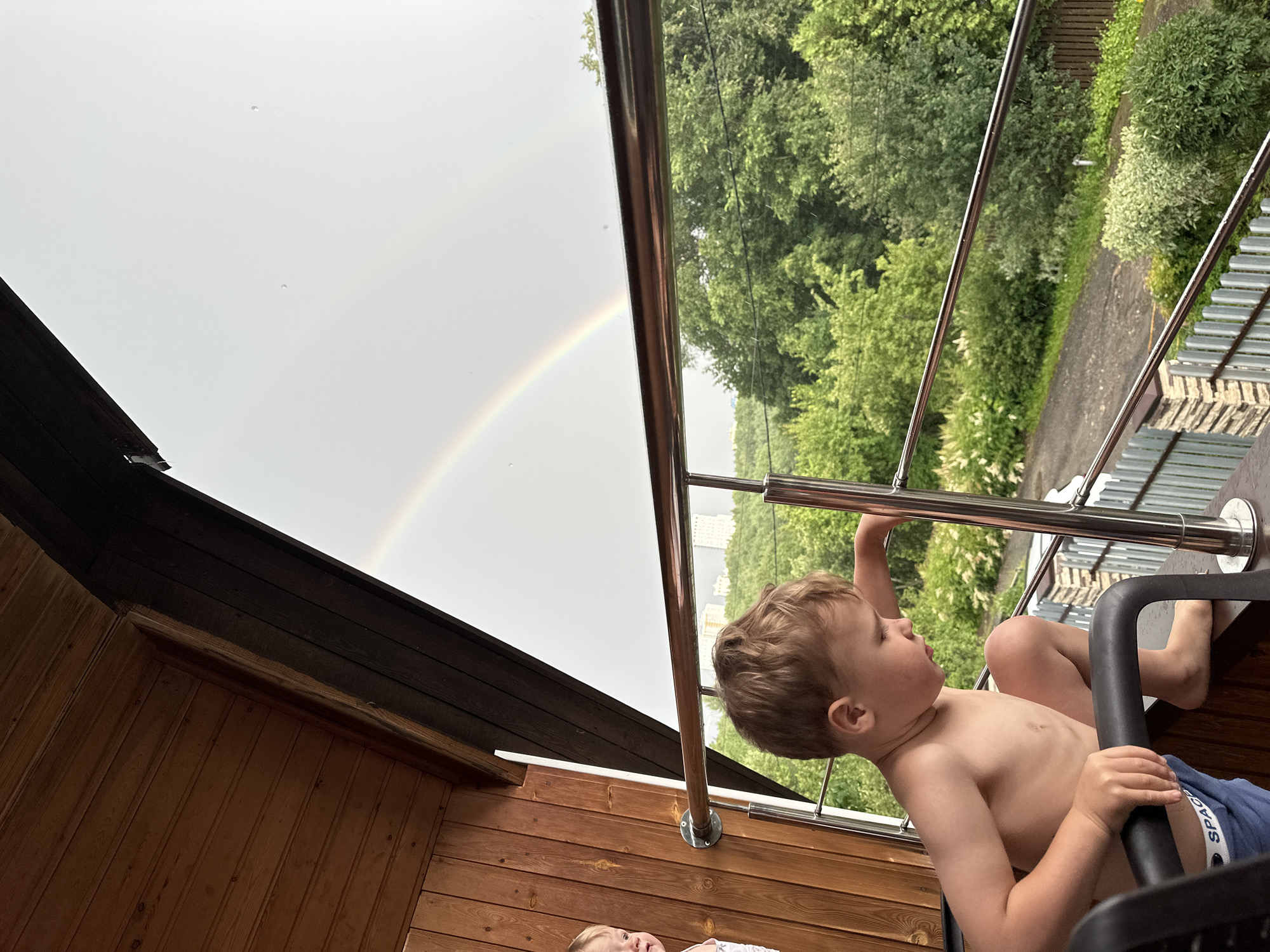 A young boy, shirtless and wearing blue shorts with "SPACE" printed on them, sits on a black chair on a wooden balcony. He is looking up at a faint rainbow in a cloudy sky, with his hands gripping a metal railing. To his left, a section of a wooden roof is visible. In the background, lush green trees line a pathway, and a glimpse of buildings can be seen in the distance. The weather appears to be overcast, possibly after a rain shower, indicated by the rainbow. The scene is set on a balcony, likely in a residential area, with a view of nature and distant urban elements. The boy's gaze suggests curiosity and wonder as he observes the natural phenomenon. The time of day is not precisely determinable, but the overcast sky and the presence of a rainbow imply it could be late morning or afternoon.