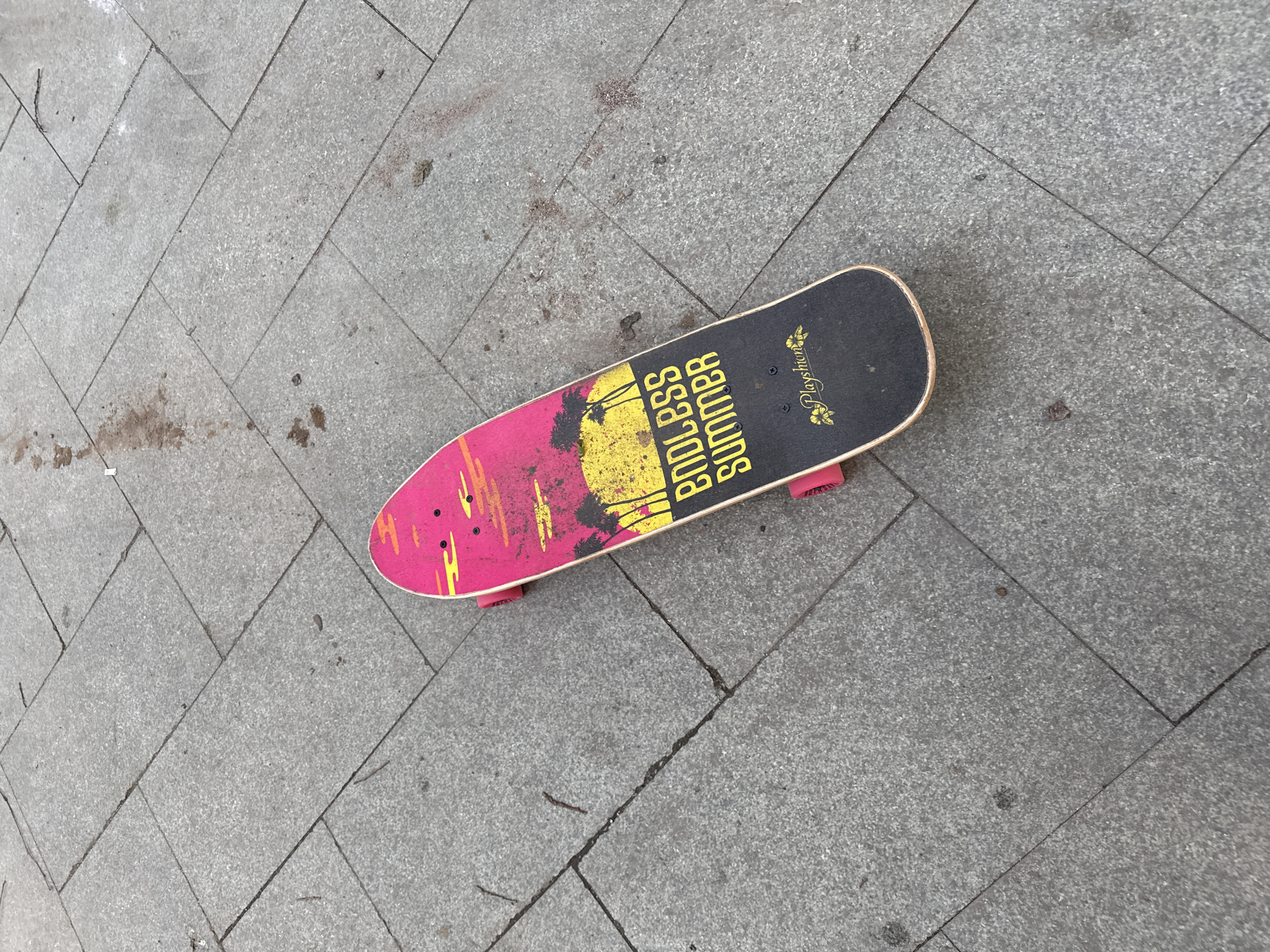 A skateboard with a pink and yellow "Endless Summer" graphic and palm trees lies on a grey stone-tiled sidewalk. The skateboard is oriented diagonally from the upper left to the lower right of the frame. The tiles are arranged in a herringbone pattern. There are small debris pieces scattered on the sidewalk. The lighting suggests it is daytime. The skateboard is the main subject, and the environment is an urban outdoor setting.