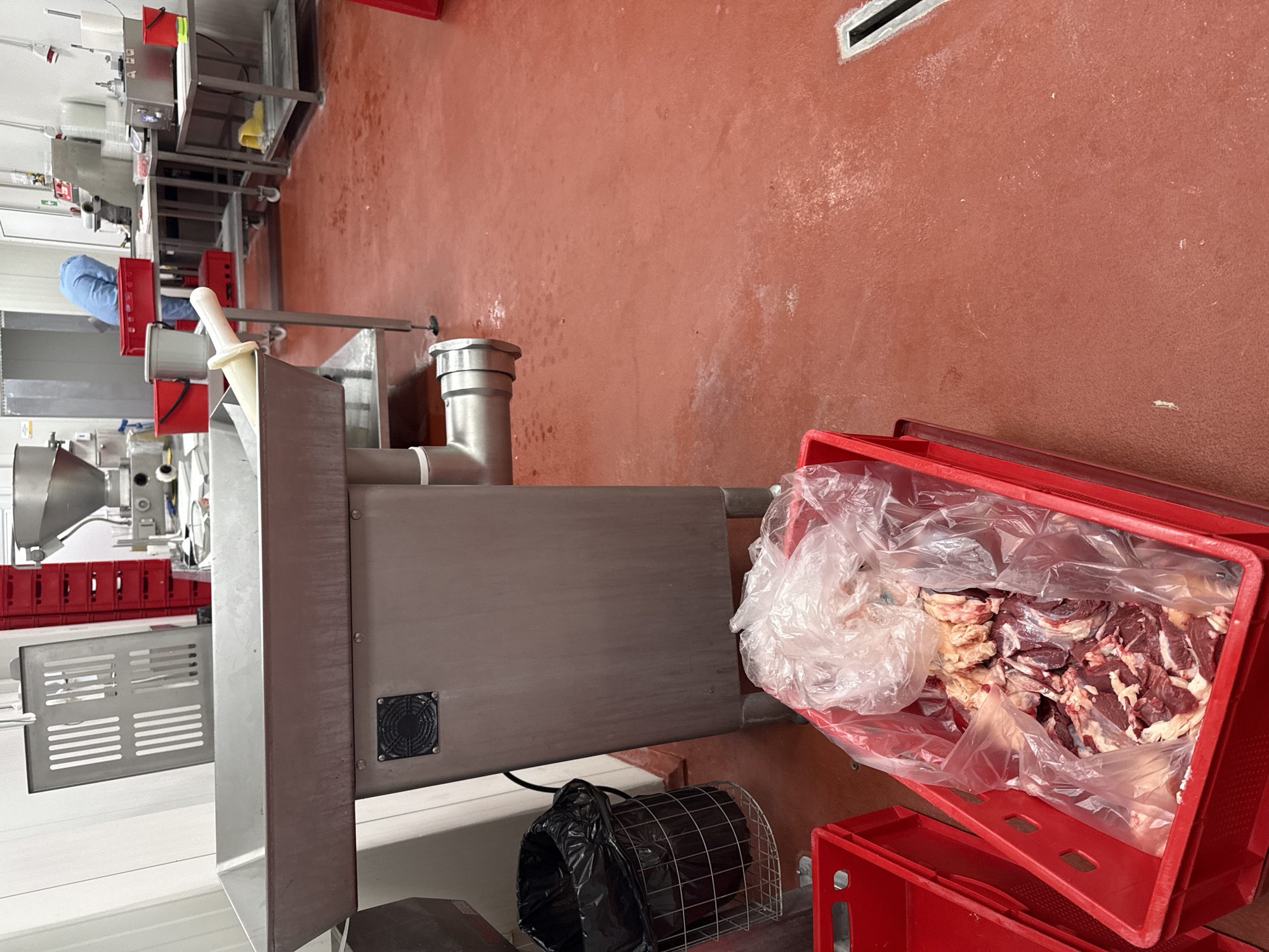 Location: Stara Zagora, Bulgaria (presumed based on the query).
Scene Description: The image presents an interior space resembling a food processing facility, possibly related to meat production. The dominant color is a reddish-brown hue, covering the floor and possibly the lower portions of the walls. The floor surface appears smooth and likely made of durable material suitable for industrial use.
Foreground: A large stainless-steel machine occupies a significant portion of the foreground. It includes a cylindrical pipe extending towards the right. A red plastic crate filled with raw meat, partially covered with a plastic bag, is located adjacent to the machine. Another red plastic crate is positioned beneath the crate holding the meat. Next to the machine, there is a metal wire basket containing a black plastic bag.
Background: In the background, there are additional pieces of processing machinery, including what looks like food grinders. A person wearing a blue uniform is partially visible near the machinery. Red plastic crates are stacked and distributed in the background. Stainless steel tables or work surfaces are also present.
Overall: The scene suggests an active food processing environment, specifically involving the handling of raw meat. The equipment and infrastructure are industrial in nature. The presence of food items and processing machinery suggests a focus on production and potential distribution.