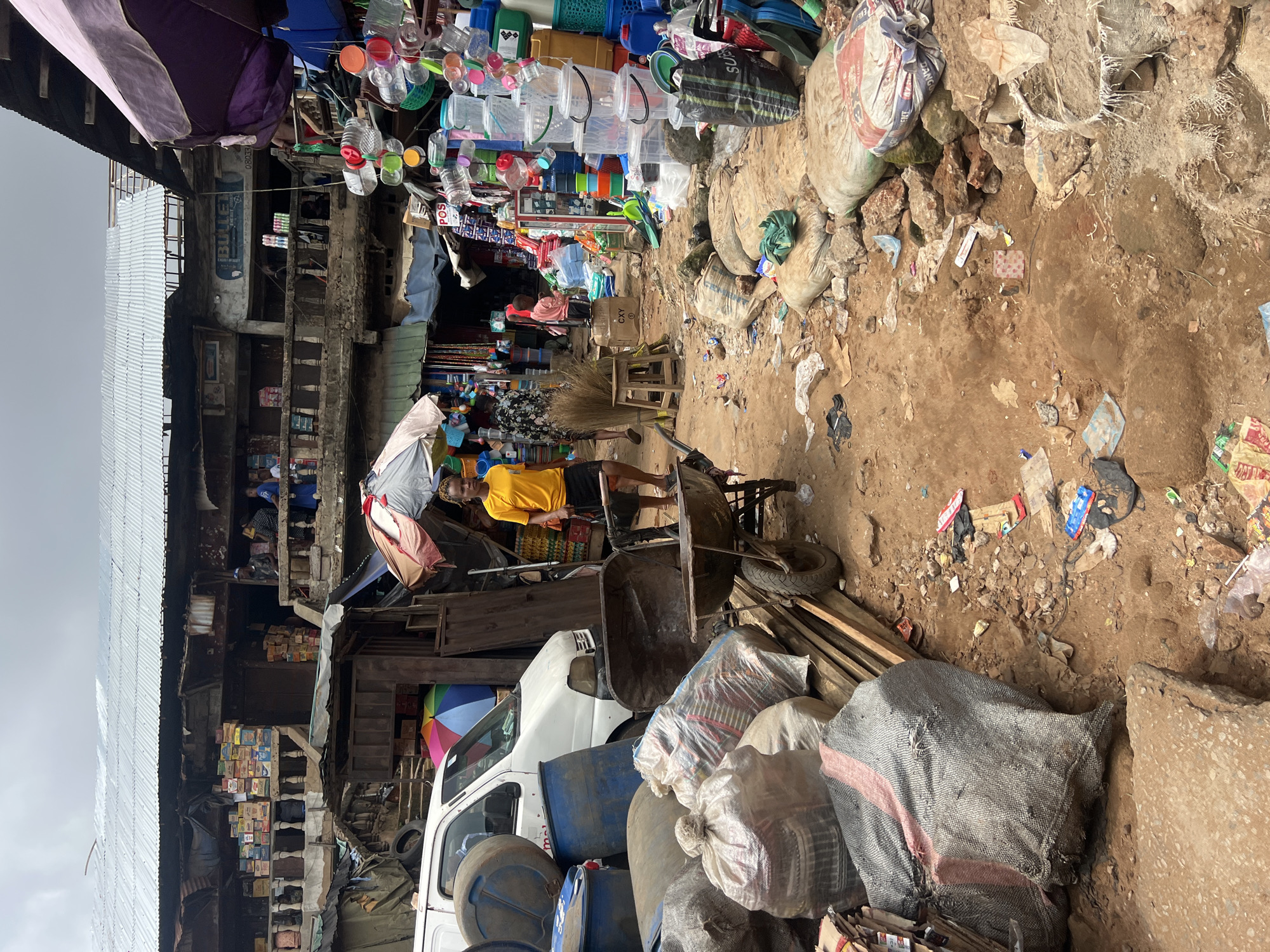 This is a media file depicting a bustling market scene, likely in Nkpor, Nigeria, characterized by an abundance of goods, people, and a somewhat disorganized setting.
The subjects include several people, predominantly engaged in market activities. One individual, wearing a yellow t-shirt and dark shorts, stands near a wheelbarrow, seemingly overseeing or participating in the transfer of goods. Another person, in a black and white patterned dress, is partially visible in the background. The market itself is the primary setting, filled with a wide array of items.
Dominating the visual field are various objects. Stacks of plastic containers, bottles, and baskets are prominently displayed, suggesting a market that sells household goods or raw materials. Sacks filled with what appear to be grains or other bulk items are piled on the ground. A white van is parked among the goods, possibly for transportation. Colorful umbrellas and tarps are also visible, providing shade or indicating stalls. The ground is a mixture of dirt and debris, highlighting the often-unpaved nature of such markets.
The activity appears to be the daily operation of a busy marketplace. People are likely buying, selling, and transporting goods. The scene conveys a sense of commerce and daily life in a bustling environment.
Notable details include the overcast sky, suggesting it could be morning or a cloudy day. The architecture in the background shows multi-story buildings with open fronts, typical of traditional markets. Some visible text includes "BULLET" on a poster and what appears to be "POS" on a sign. The overall impression is one of vibrant but somewhat chaotic market activity, common in many parts of Nigeria.