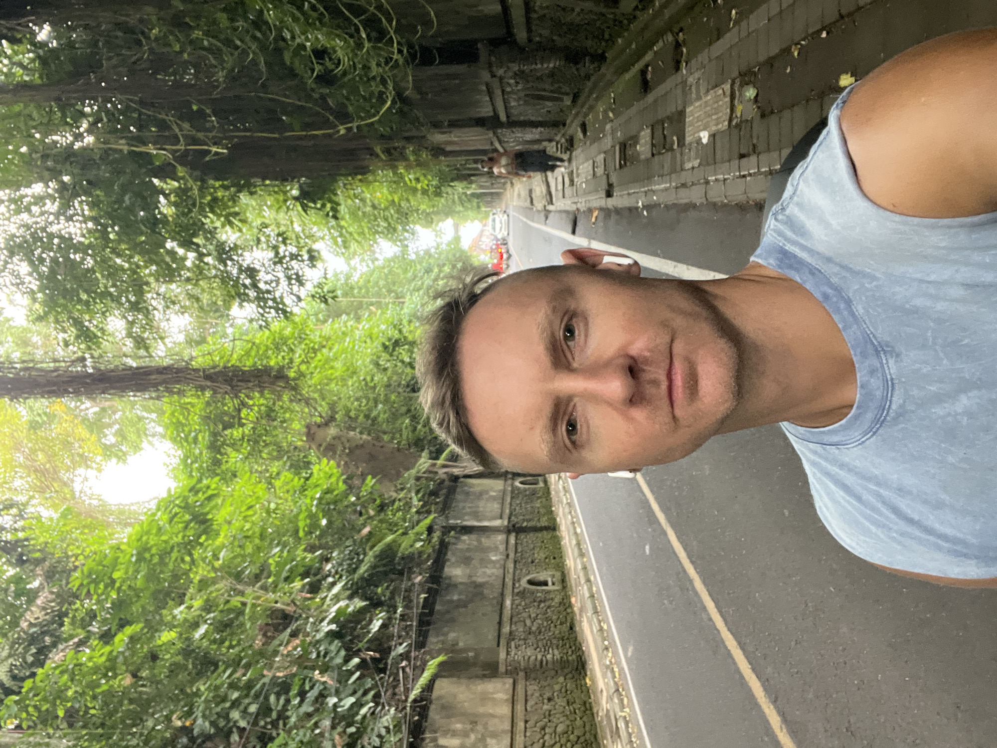 The photograph shows a man in a blue shirt on a road lined with dense trees and stone walls in Ubud District, Indonesia. The road has a yellow dividing line, and a figure can be seen walking in the distance. Cars are also visible further down the road. The environment is lush with greenery.