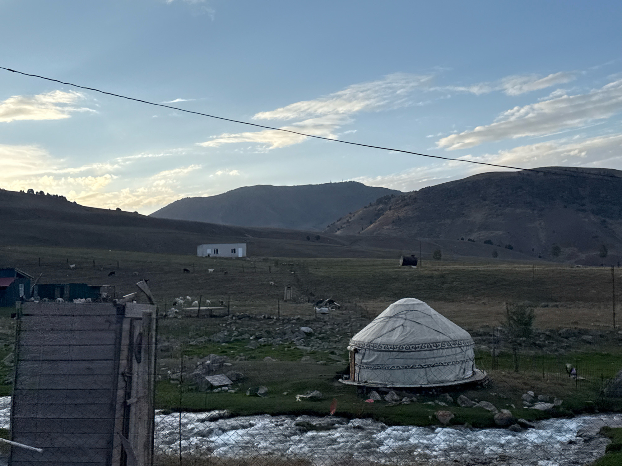The image depicts a rural landscape in Chunkurchak, Kyrgyzstan, featuring a traditional yurt situated near a fast-flowing river. The yurt, a circular tent structure with a conical roof covered in a white fabric, is the central subject in the foreground on the right. The surrounding terrain is hilly and covered in dry grass, with scattered rocks. In the middle ground, several goats are visible grazing in a fenced area, and a simple white building with windows is present. To the left, a portion of a wooden structure, possibly part of a fence or outbuilding, is in the immediate foreground. The background consists of rolling, dark hills under a pale blue sky with wispy clouds. A thin wire stretches across the upper portion of the image. The overall scene suggests a remote, pastoral setting during daylight.
