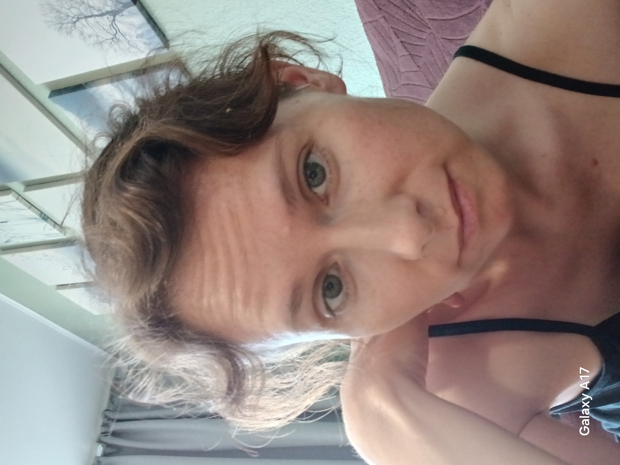 Here is a description of the image:
A selfie shows a light-skinned woman with light brown hair and hazel eyes. She is wearing a black tank top. Her hair is wavy and slightly messy. There are framed images on the wall behind her. There is also a lavender-colored textured material that looks like a blanket. A grey curtain is partially visible. The photo was taken using a Galaxy A17 smartphone. There is nothing in the image indicating it was taken in Yekaterinburg, Russia.