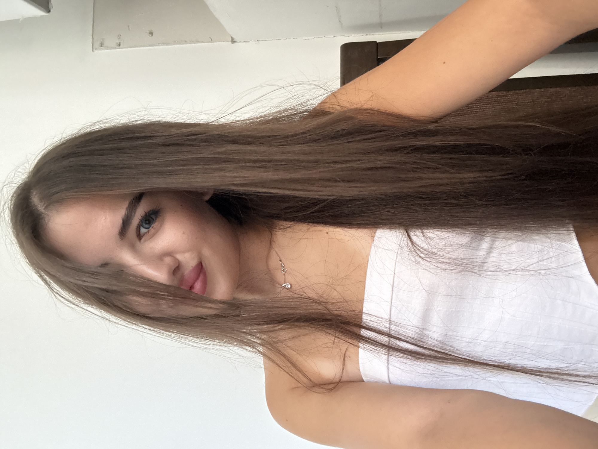 A young woman with long brown hair is captured in a close-up selfie. Her hair partially obscures her face, with strands falling across her left eye and cheek. She has blue eyes and is wearing a white strapless top. A delicate necklace with a small pendant is visible on her neck. The background is a plain white wall. The lighting suggests an indoor setting, possibly a home or studio, with a hint of a wooden chair visible on the right side of the frame. The image does not provide enough information to determine its specific location in Chișinău, Moldova.