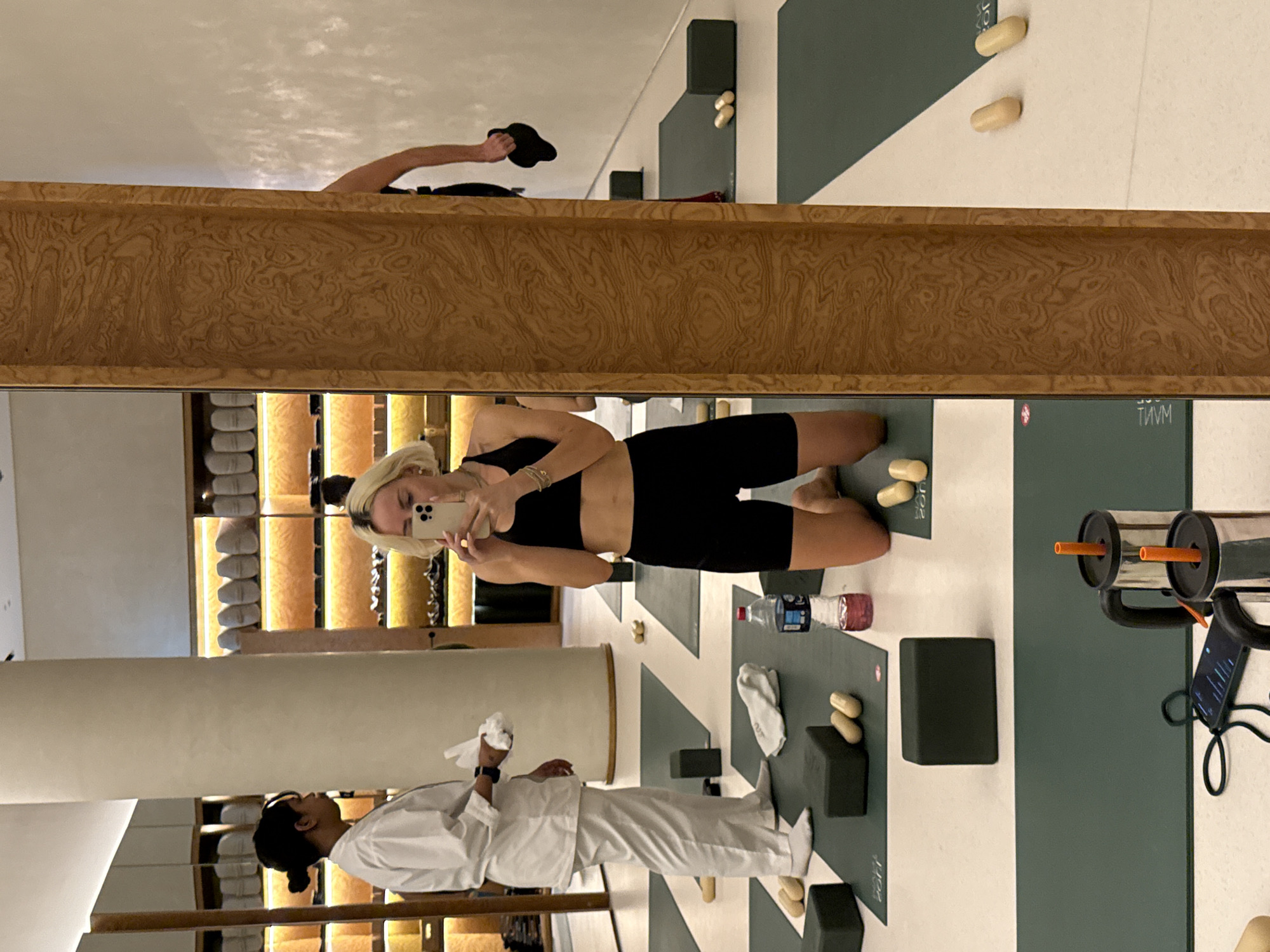 A woman is taking a selfie in a mirror in a fitness facility in Dubai, United Arab Emirates. A person wearing a white uniform is walking along a yoga mat. The environment includes a mirrored wall, towels on shelves, water bottles, yoga mats, blocks, and exercise equipment.