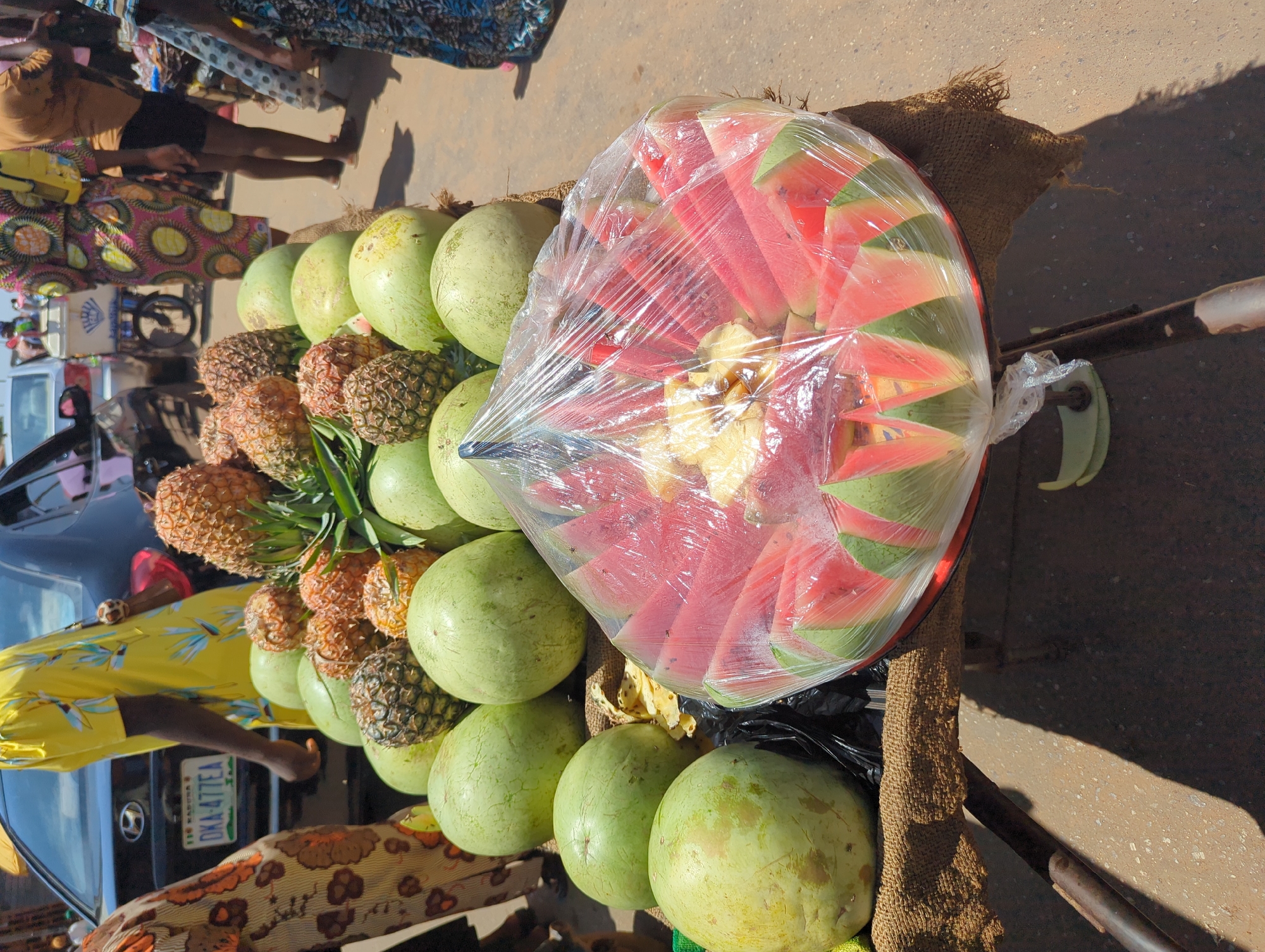 The image depicts a street market scene in Narayi, Nigeria, characterized by a fruit vendor's pushcart in the foreground and various individuals and vehicles in the background.
The foreground is dominated by a metal pushcart, its top covered with a coarse, light brown woven material, likely burlap. Piled on this cart are numerous whole, round watermelons, primarily light green with some yellowish patches. Interspersed among the watermelons are several whole pineapples, displaying rough, segmented, yellowish-brown to darker brown skins and green leafy crowns. On the right side of the cart, a round, red-rimmed tray holds an arrangement of pre-cut, triangular watermelon slices and irregular pineapple chunks. This tray is sealed with clear plastic wrap, which reflects light, protecting the exposed red watermelon flesh and yellow pineapple. A crumpled black plastic bag is visible beneath the tray.
In the mid-ground, behind the fruit cart, several people are visible. On the far left, a person's lower body and arm are seen, clad in a brown and orange patterned fabric. Towards the center-left, an individual wearing a bright yellow shirt with a blue and green floral pattern walks away from the viewer. Behind this person, a dark-colored Hyundai sedan is parked or moving slowly; its rear license plate, "DKA-477EA," along with "NIGERIA" and "F.C.T ABUJA," is clearly legible. Further to the right, another person wears a brightly patterned garment featuring pink, yellow, and green circular motifs. Additional pedestrians are distributed across the background, some in colorful attire. A white pushcart with "BELVEDERE" partially visible on its side is being operated by an individual further down the street. A light-colored vehicle is also visible in the distance.
The ground surface is a reddish-brown, uneven terrain, consistent with an unpaved or poorly paved road. The scene is brightly illuminated by direct sunlight, casting distinct shadows from the cart and people, indicative of a daytime setting. The collective elements signify an active commercial street environment within Narayi.